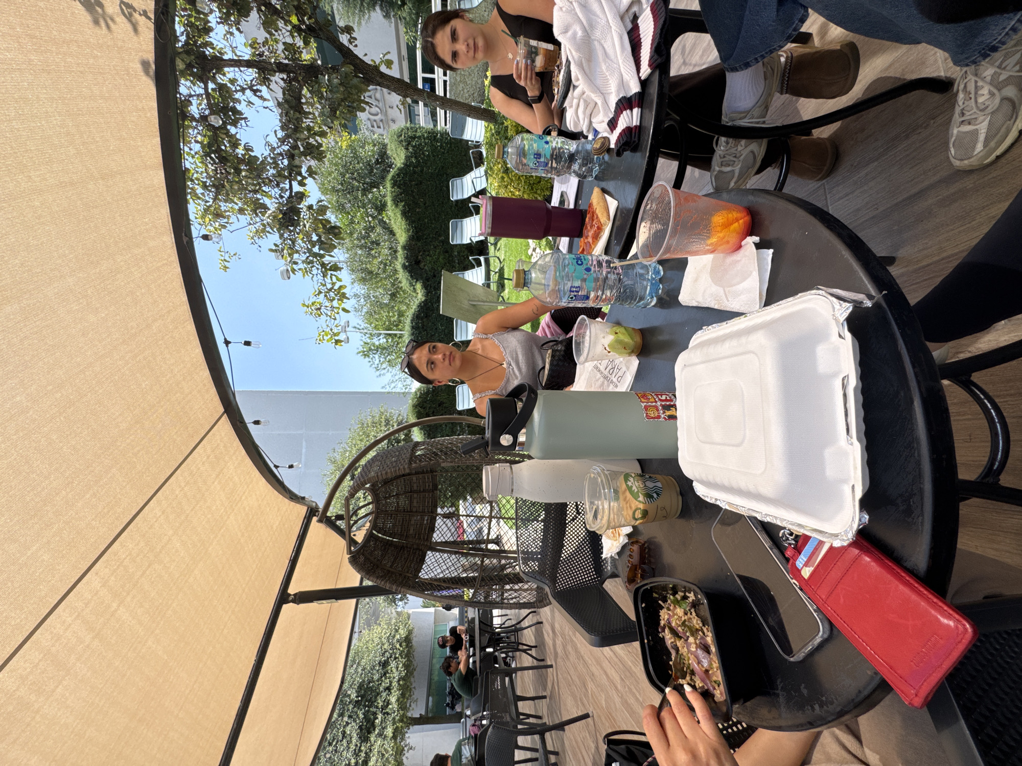 The image depicts an outdoor scene, likely a cafe or patio, with multiple people seated at tables. Two women are visible in the foreground at one table, and another woman is seated at a table further back. The tables are covered with food containers, water bottles, and other personal items such as a red wallet and a phone. One of the women in the foreground is looking towards the camera. Other individuals are visible in the background seated at various tables. The location appears to be outdoors under a large awning with trees and greenery visible in the background. Based on the buildings and vegetation, the location is consistent with an outdoor patio or cafe in Heroica Puebla de Zaragoza, Mexico.