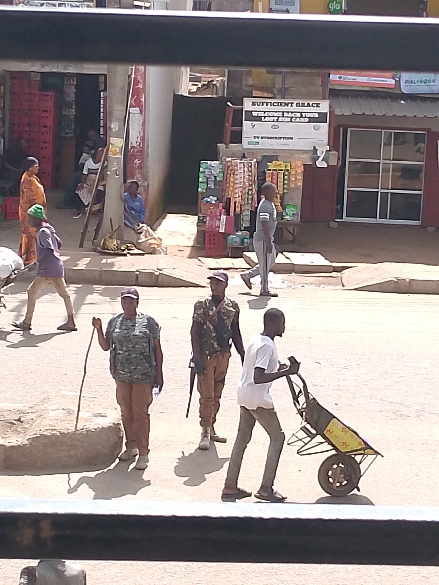 This media file shows a street scene in Bukuru, Nigeria, likely during the daytime, given the bright sunlight and shadows. The foreground features several individuals, including two people in what appear to be security or military uniforms, one holding a rifle and the other a stick. A third person is pushing a wheelbarrow. In the background, more people are visible, some near shop entrances or standing on the street.
The setting appears to be a busy commercial area with shops lining the street. One shop has a sign that reads "SUFFICIENT GRACE WELCOME BACK YOUR LOST SIM CARD" along with "TV SUBSCRIPTION." Another sign above it indicates "glo," a telecommunications company. Red crates are stacked near one of the shop entrances. The ground is paved and appears somewhat dusty or uneven. The overall impression is of a typical day in a Nigerian market or business district. The emotions of the people are not clearly discernible due to the distance and image quality, but the scene suggests normal daily activity.
