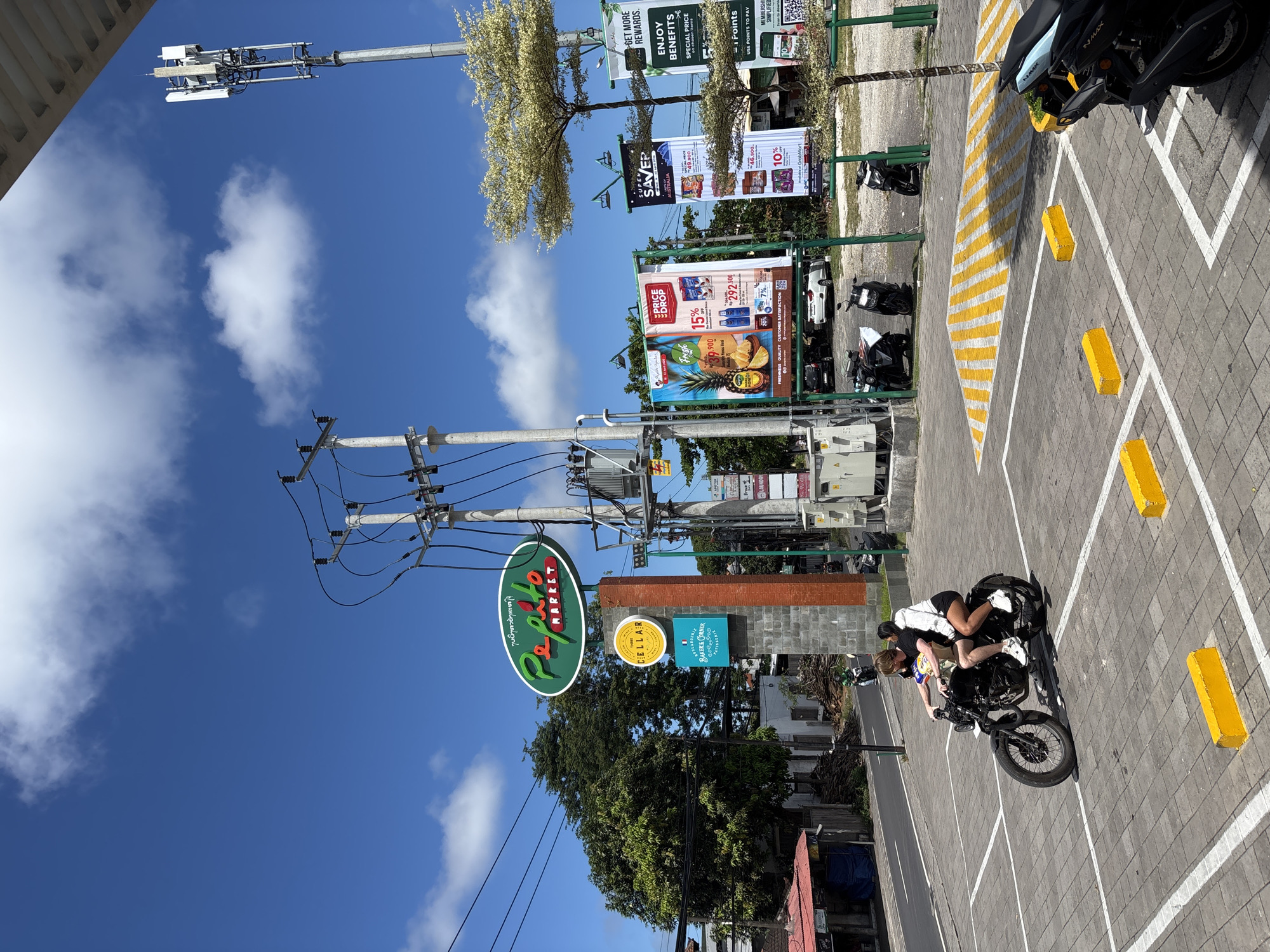 The image depicts a bustling outdoor commercial area in Kuta Selatan, Indonesia, under a bright, clear blue sky with scattered white clouds. The scene primarily features a paved parking lot made of grey bricks, marked with white and yellow lines and yellow concrete parking stoppers.
In the foreground, two individuals are on a dark-colored motorcycle, parked in a designated space. The driver, wearing a white t-shirt and dark shorts, is holding a yellow can, while a passenger is seated behind them, also in a white t-shirt and dark shorts. Other motorcycles and scooters, including a Yamaha NMAX, are parked further in the lot.
The background is dominated by numerous commercial signs and banners. A prominent green oval sign reads "Pepito MARKET" in white and red lettering, with smaller signs for "THREE CELLAR" and "BAKER'S CORNER PATISSERIE" below it. Large promotional banners advertise "SUPER SAVER AUSTRALIA," "PRICE DROP 15% OFF," and "Fresh" produce priced at "39.900" and "292.500." Other banners display text such as "GET MORE REWARDS.," "ENJOY BENEFITS.," "SPECIAL PRICE," "i-POINTS," "USE POINTS TO PAY," and "MEMBERSHIP SIMPLIFIED," with one banner mentioning "GlobMart."
Utility poles with power lines and cellular antennas rise against the sky. Several green trees with light-colored foliage and parts of buildings, including a red brick wall, are visible. The strong sunlight casts distinct shadows, indicating midday or early afternoon. The overall scene suggests a vibrant retail environment.