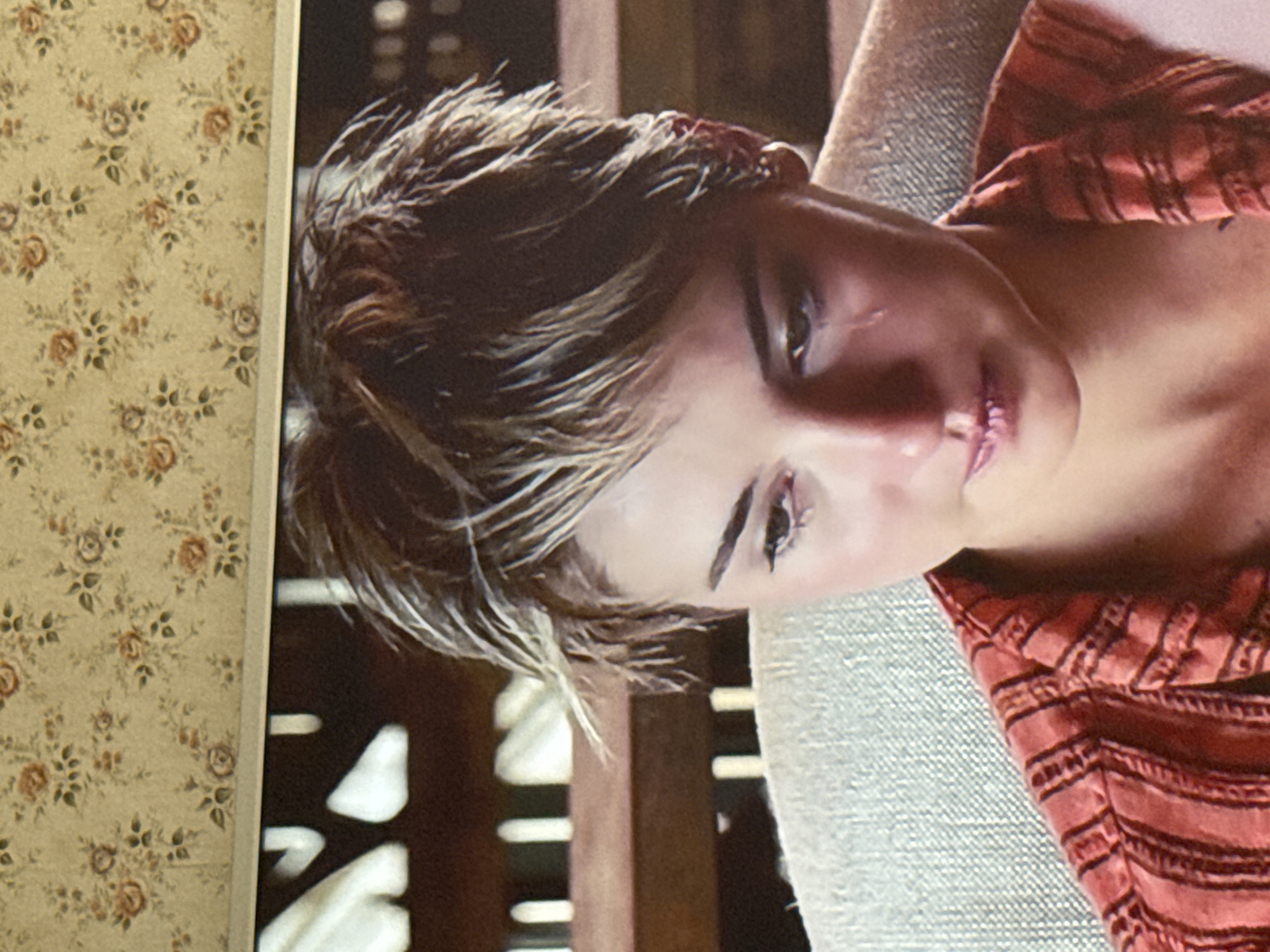 A close-up, slightly angled photograph depicts a person with short, light brown hair with blonde highlights. The individual is fair-skinned, with visible eyelashes, and a slight scar near their right eye. They are wearing a red and brown striped garment. The person is leaning against a light gray textured surface. Behind them, a dark wooden structure is visible. To the left of the structure is a framed print with a floral design of small roses on a beige background. The location, Mytishchi, Russia, cannot be verified from the image.