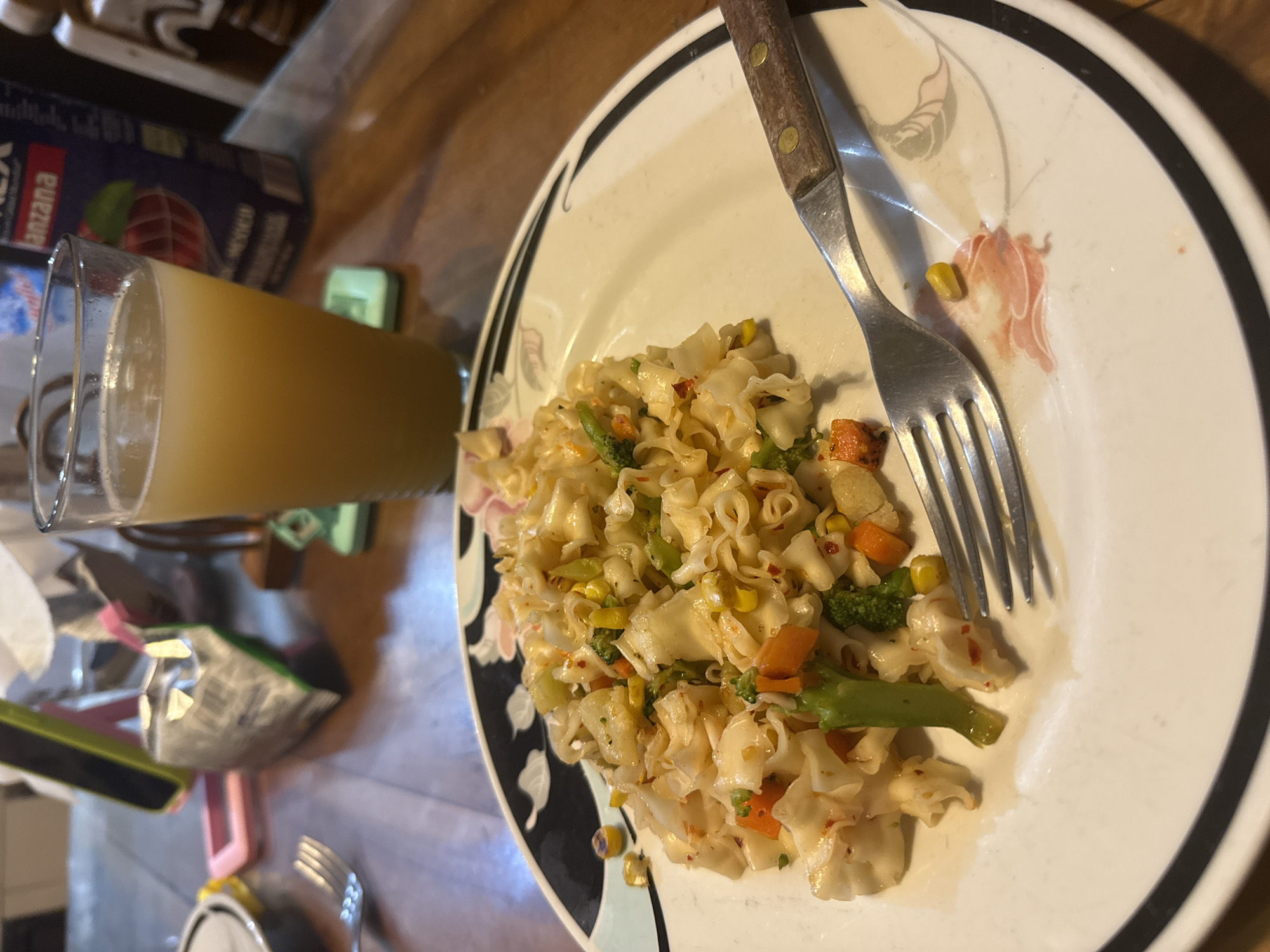 A plate of pasta salad with vegetables and a fork rests on a table. Next to the plate is a glass of orange juice and a dark blue juice box. The location is likely in Xochitepec, Mexico, due to the packaging of the juice box. The meal consists of pasta salad with visible components like corn, carrots, and broccoli. The environment appears to be indoors on a wood-grain surface.