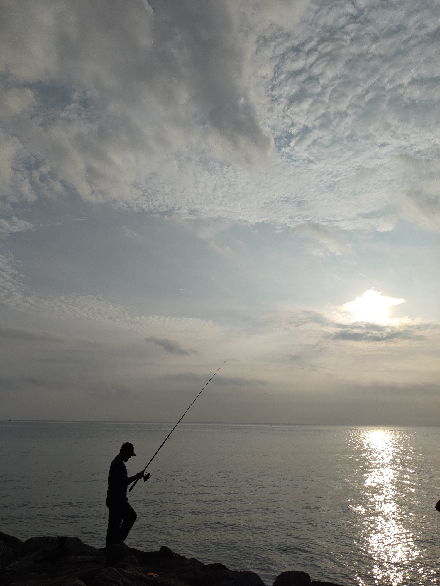 The image presents a serene coastal scene captured at either sunrise or sunset in Lhokseumawe, Indonesia. A lone angler, silhouetted against the bright sky, stands on dark rocks at the water's edge. The individual, appearing to be an adult male wearing a cap, holds a fishing rod with a reel, seemingly engaged in casting or waiting for a catch.
The vast expanse of the sea stretches calmly to the horizon. A prominent, low-lying sun, partially obscured by clouds, creates a brilliant, shimmering path of light across the water's surface, reflecting intensely towards the foreground. The sky above is largely covered with various cloud formations, ranging from scattered, textured clouds in the upper left to more diffused, layered clouds across the rest of the firmament, contributing to the soft, atmospheric lighting.
The overall impression is one of peaceful solitude and patience, highlighting the quiet activity of fishing amidst a beautiful natural backdrop. No visible text is present in the image.