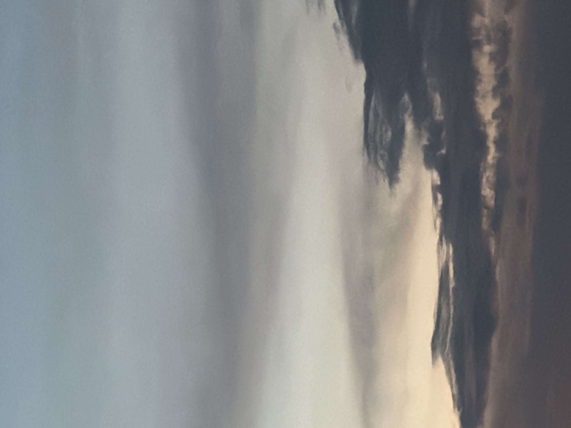 A vertical, full-frame, close-up shot captures a gradient of twilight sky transitioning from a light, pale blue at the top to darker, muted tones of gray and purple at the bottom. The sky is filled with layers of clouds. The upper half of the image shows wispy, diffused clouds that blend into the clear sky. As the view descends, the clouds become denser and more defined, with darker gray formations dominating the lower half. Some of these darker clouds have a textured, almost stormy appearance, while others are softer and more shadowed. The very bottom edge of the frame shows the slightest hint of a lighter, warm hue, possibly from a distant horizon or reflected light. The image appears to be taken from an open area, such as a park or a viewpoint, in Chaikovsky, Russia, during the twilight hours. There are no people, identifiable landmarks, or distinct objects visible in the frame; the focus is solely on the atmospheric conditions of the sky.