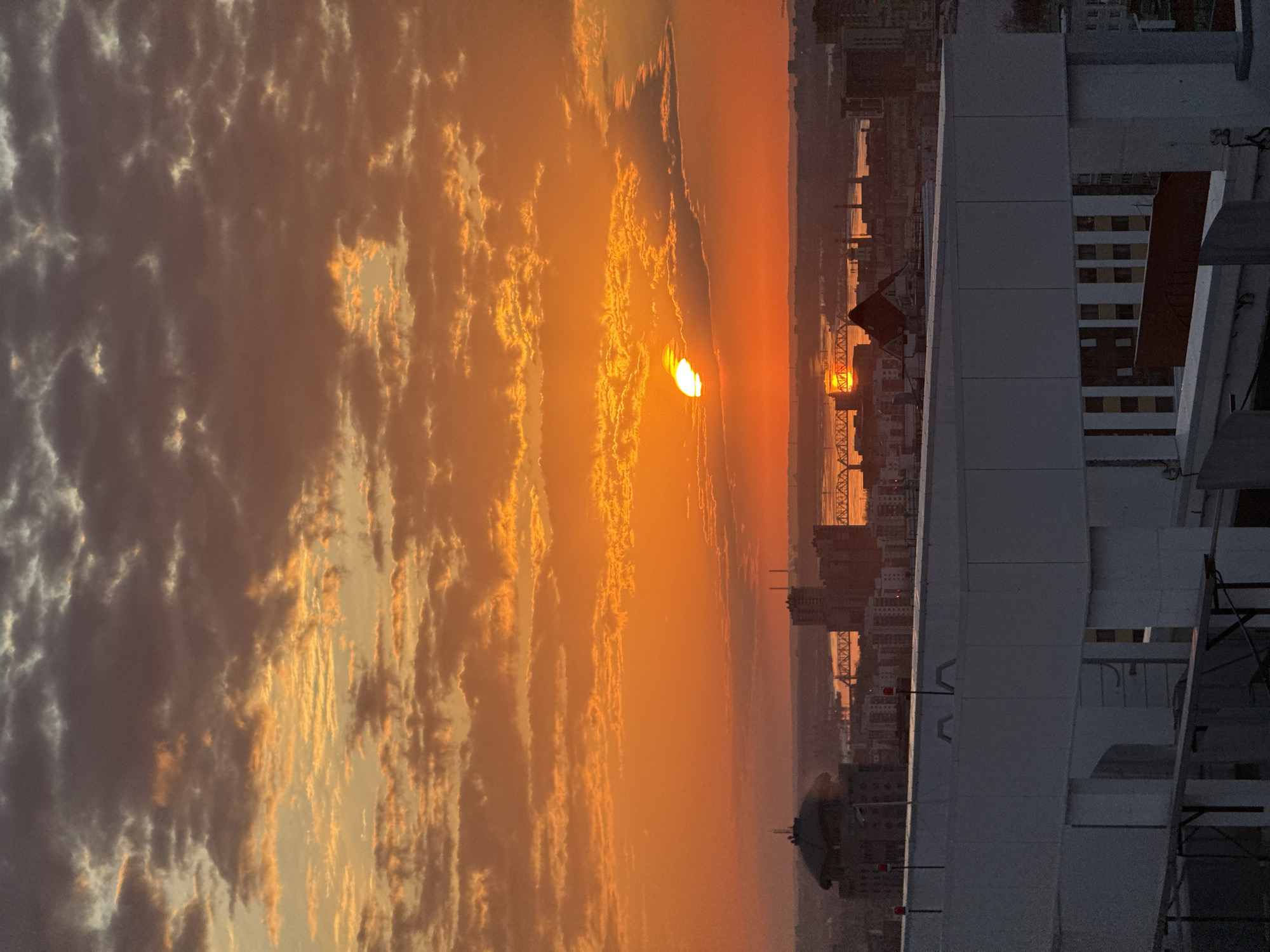 The image depicts a sunset in Perm, Russia, as viewed from a high vantage point, likely a tall building or elevated area. The sky is dominated by orange and yellow hues, with scattered clouds. The setting sun is partially obscured by the clouds, but its intense light is still visible. In the distance, the skyline of Perm is visible, with the Kama River running through it. A bridge is also visible. In the foreground, a portion of a building is present, featuring white and gray surfaces. The overall scene captures the urban environment bathed in the warm light of the setting sun.