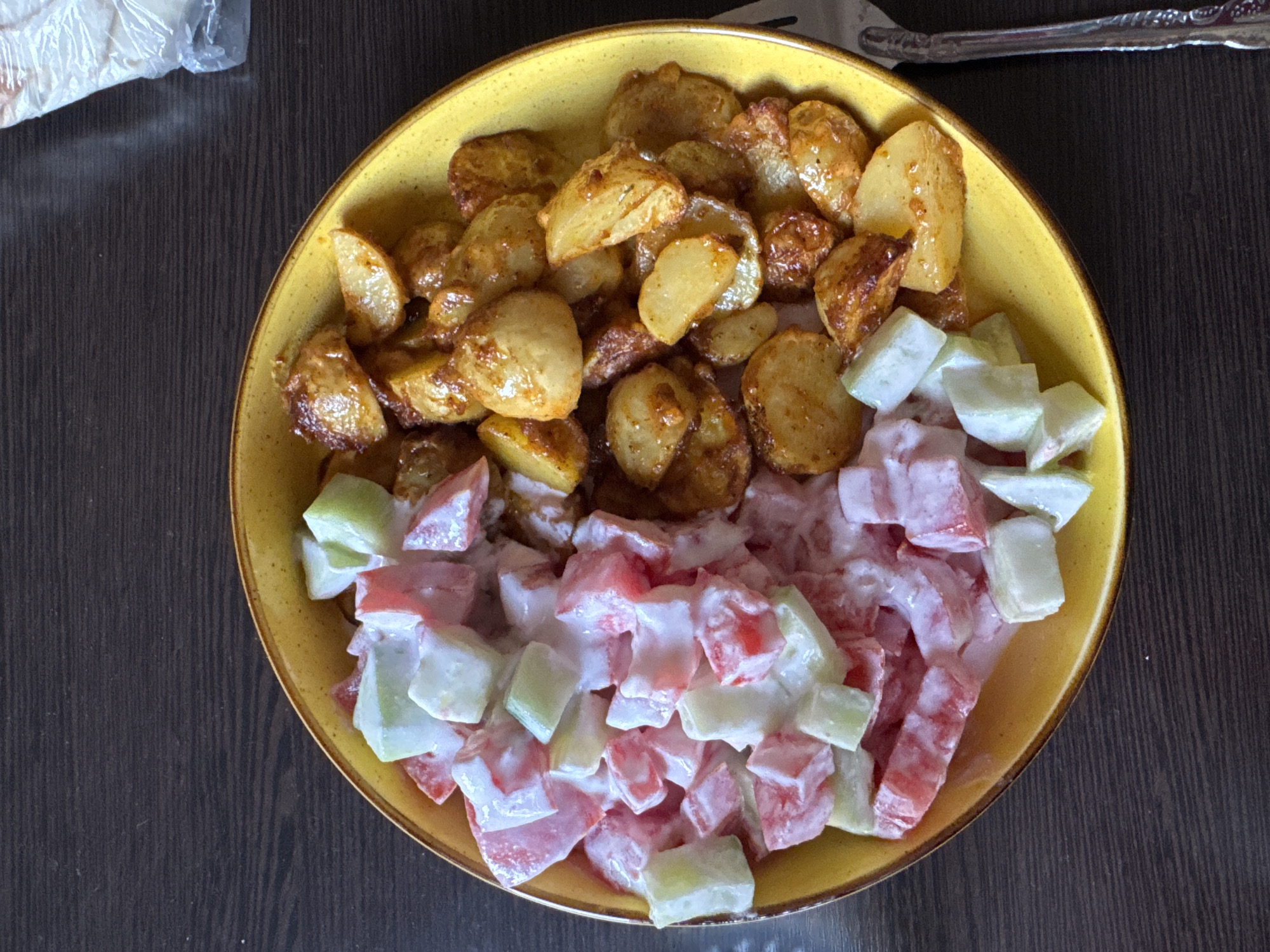 A close-up, top-down shot shows a yellow bowl filled with food on a dark wooden surface. The bowl is divided into two sections. The left side contains roasted potato wedges that are golden brown and appear to be seasoned. The right side of the bowl holds a salad made of diced tomatoes and cucumbers, mixed with a creamy white dressing, possibly mayonnaise or yogurt. The overall presentation suggests a meal, likely lunch or dinner, served in a casual setting. A fork with an ornate handle is visible at the top of the frame, resting on the wooden surface. In the upper left corner, a portion of a plastic-wrapped item, possibly bread or another food item, is also visible. The lighting suggests an indoor environment, and there are no strong indications of the time of day or weather conditions. The image is centered on the food, with the wooden table and the edge of a fork providing context. There is no visible text in the image. The scene is set in Yerevan, Armenia.