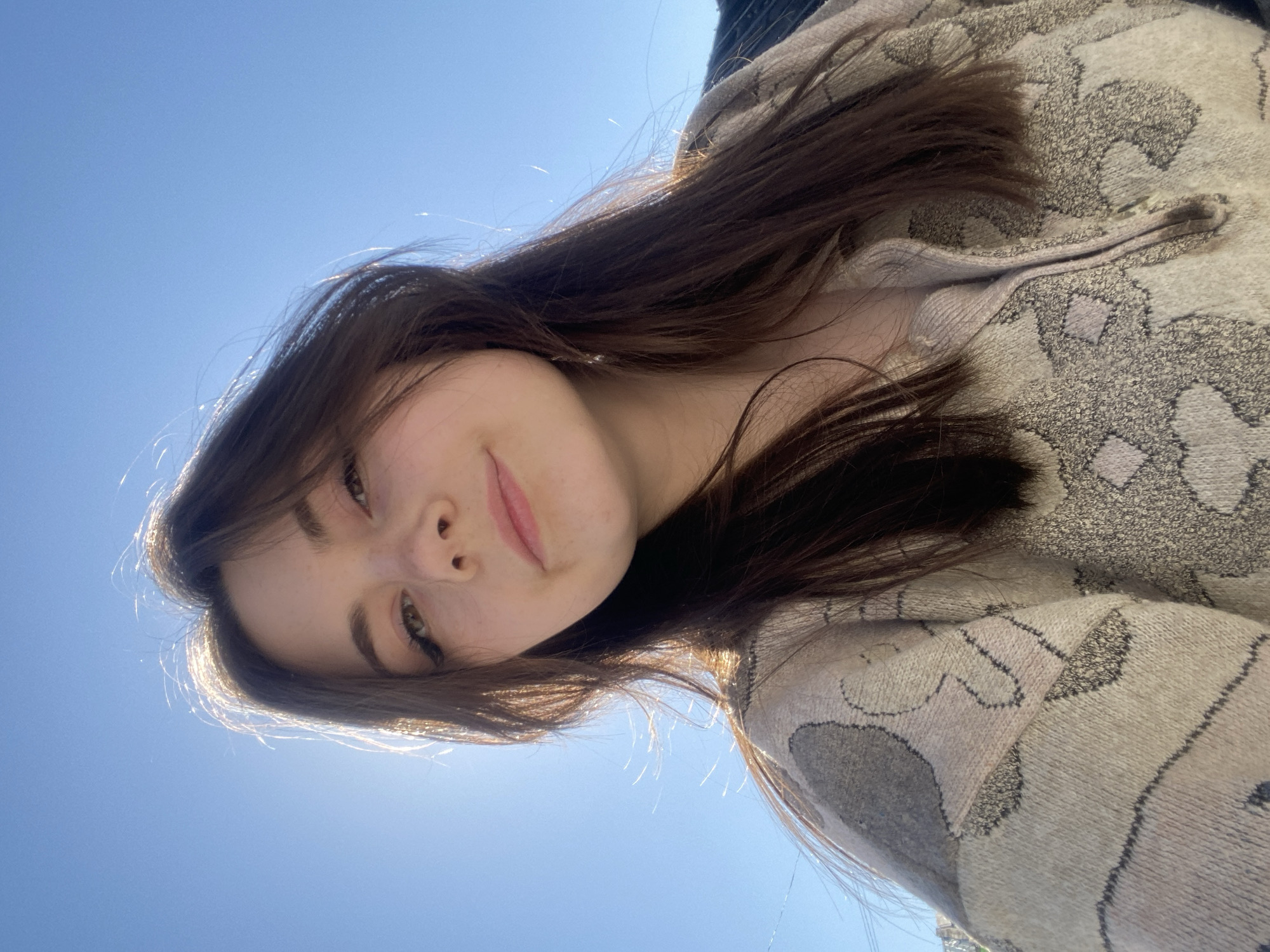 A young woman takes a selfie from a low angle under a bright blue sky, perhaps in the town of Sosnovo, Russia. She has light skin, brown hair with bangs, and is wearing a light-colored patterned sweater. The focus is on her face, with a slight smile, and the clear sky is visible in the background.