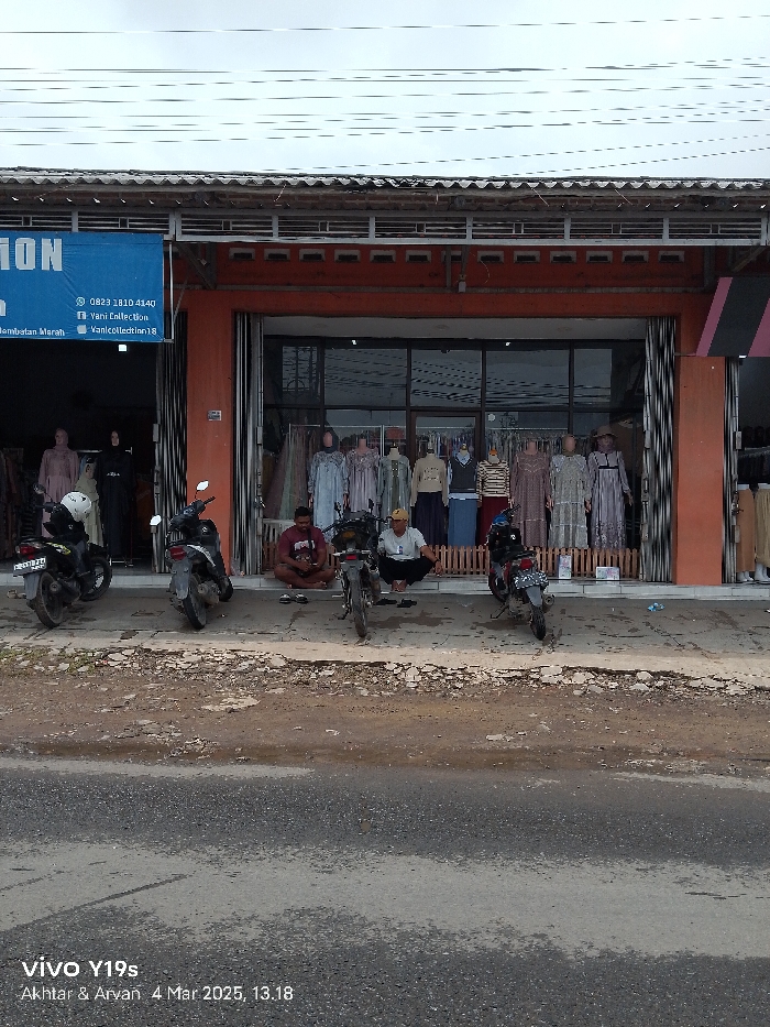The image shows a storefront located at latitude -6.468 and longitude 108.297 in the city of Jatibarang, Indonesia. The storefront has a large glass window displaying various clothing items on mannequins. Two men are sitting outside the store on the sidewalk. One man is wearing a red shirt and the other a light-colored shirt. Three motorbikes are parked in front of the store. The storefront is painted orange and has a blue sign above the door with the phone number 0823 3810 4140 and the name “Yani Collection” written on it. The sign also mentions a social media account: “vanicollection18”. The sidewalk in front of the store is made of concrete and is cracked in places. The street in front of the storefront is paved with asphalt. The sky is overcast and gray.