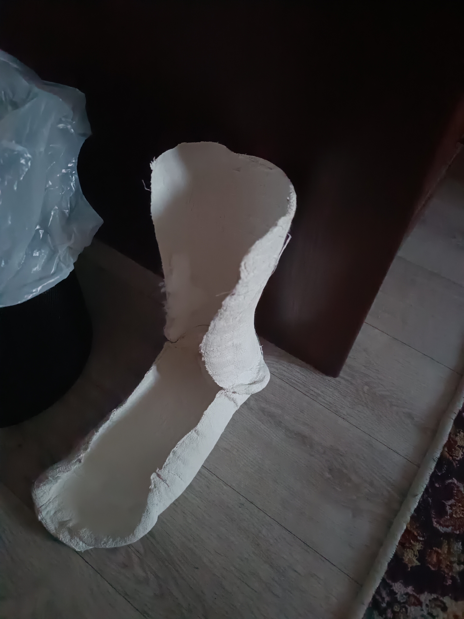 The image displays a white plaster cast of a foot and lower leg, lying on a light-colored wooden floor. The cast is positioned diagonally, with the heel towards the foreground and the upper part extending towards the background. To the left of the cast, a crumpled white plastic bag is visible, partially obscuring a black cylindrical object. Behind the cast, a dark brown, possibly wooden, surface is present, suggesting furniture or a wall. In the lower right corner, a portion of a rug with a dark background and intricate floral patterns in red, yellow, and blue is visible. The lighting in the image is dim, creating shadows and highlighting the texture of the plaster. The overall impression is of an object placed in a domestic or storage setting, possibly discarded or awaiting further use. There is no visible text in the image. The context of Taraz city administration, Kazakhstan, is provided as external information and cannot be inferred from the visual content of the image itself.