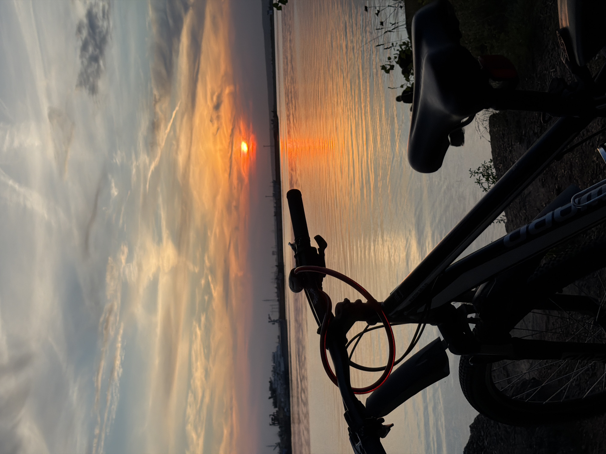 This is a vertical, medium-shot, close-up photograph of a bicycle parked by a body of water at sunset. The bicycle, predominantly in silhouette, is positioned in the foreground and takes up the majority of the left and bottom of the frame. The handlebars, seat, and front wheel are clearly visible. A red cable lock is looped around the handlebars.
The background features a wide expanse of water reflecting the warm hues of the sunset. The sun itself is visible near the horizon, casting an orange glow across the sky and water. In the distance, along the far bank of the water, are the silhouettes of industrial structures, including cranes and buildings, suggesting an industrial area. The sky above is filled with wispy clouds, tinged with orange, pink, and blue.
The scene is likely located near the Kama River in Chaikovsky, Russia, given the presence of industrial infrastructure typical of a port city on a large river. The overall mood is serene and peaceful, characteristic of a quiet evening by the water. There is one small, silhouetted figure visible in the distance, sitting by the water's edge, adding a sense of scale and solitude to the landscape. The lighting conditions indicate that it is late evening, with the sun setting. The ground in the immediate foreground appears to be rocky and uneven.