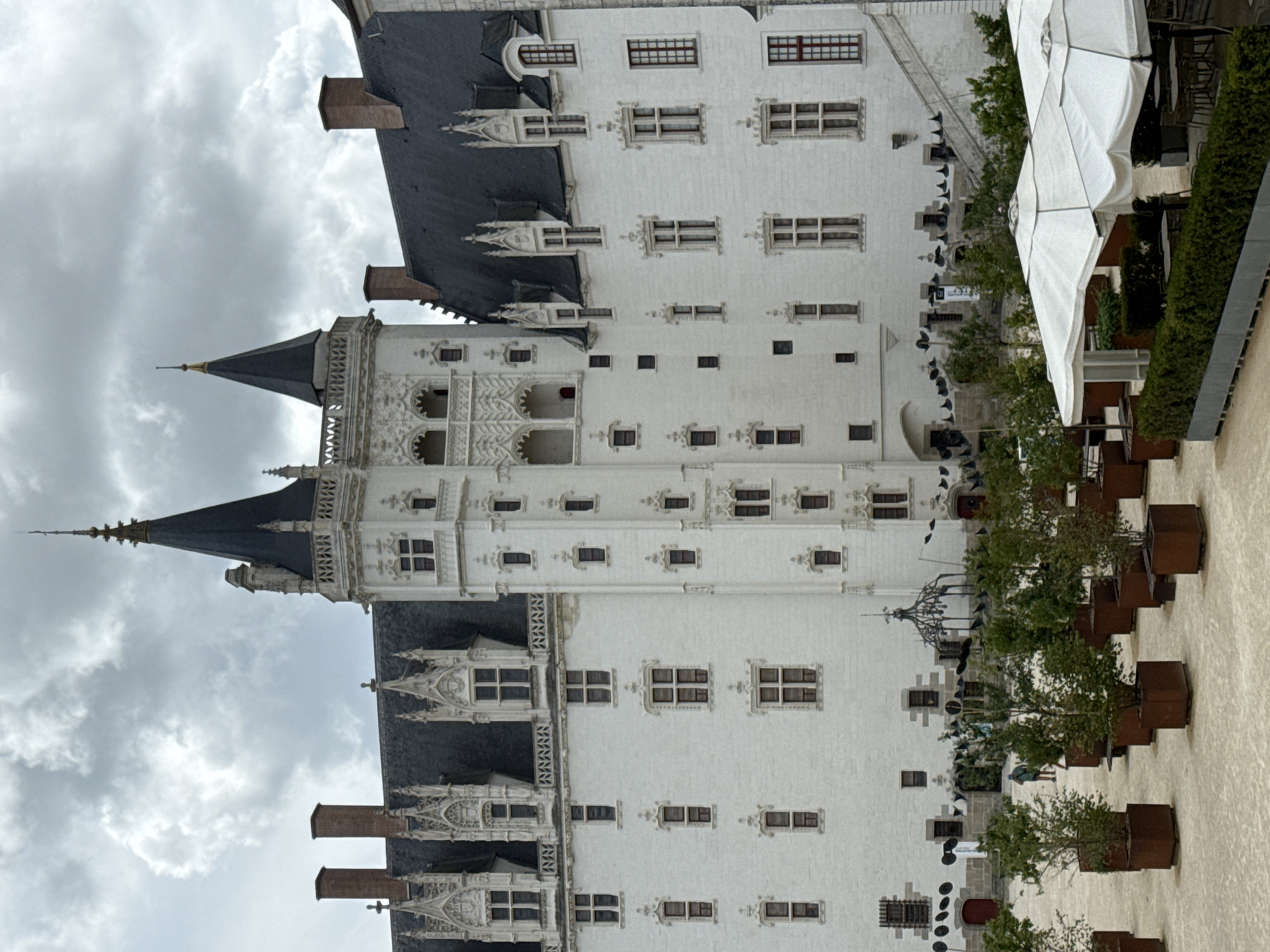 This image depicts the Château des ducs de Bretagne in Nantes, France. The imposing white stone castle features dark, pointed roofs and multiple towers, with a central tall tower dominating the skyline. Ornate architectural details, including decorative window frames and battlements, are visible on the castle's facade. In the foreground, a courtyard is furnished with several large white umbrellas providing shade over outdoor seating areas. Rows of potted plants, some appearing to be young trees, are arranged in weathered metal planters. A person is visible in the middle ground walking across the courtyard. The sky is overcast with visible clouds, suggesting a temperate climate.