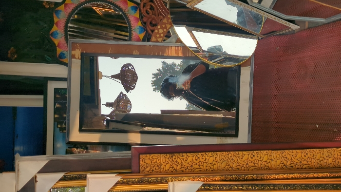 A vertical image depicts a self-portrait captured through the reflection in a rectangular mirror within what appears to be a mirror and frame shop in an urban area of Dhaka, Bangladesh.
The central subject is a person reflected in a large, upright rectangular mirror. The individual is holding a light-colored smartphone with their right hand, positioned to take the photograph, obscuring part of their face due to glare and the device. They are wearing dark-colored clothing, possibly a shirt or jacket, and have a dark, voluminous mass around their head, which could be hair or a head covering. Behind the person in the reflection, an outdoor scene with bright sky and green foliage from trees is visible, suggesting an open storefront or window. Two woven, bell-shaped pendant lights are reflected hanging above the person's head within the mirror.
To the left, stacked ornate picture frames are visible, featuring intricate gold-colored scrollwork patterns on maroon borders. To the upper right, a round mirror with a colorful, decorative frame displaying blue, pink, and yellow petal-like designs is partially visible. Below this, towards the bottom right, several smaller, unframed or thinly framed mirrors are propped up, reflecting distorted portions of the environment. The ground in the foreground is covered with a dark red, textured, possibly rubber mat with a repeating dimpled pattern. In the background, behind the main mirror, framed artworks depicting a dark blue water scene and city lights are partially visible.