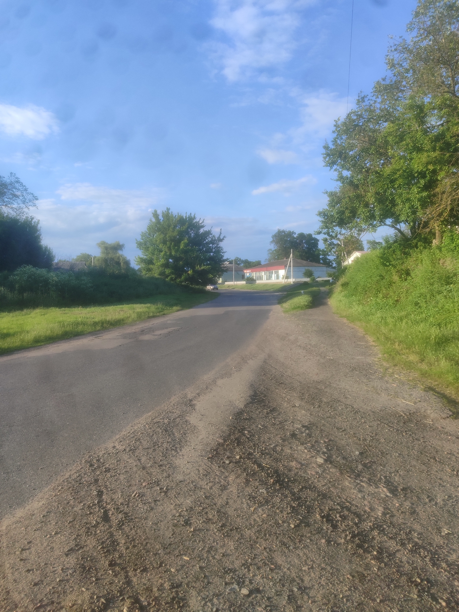 An asphalt road runs through a rural area with a clear blue sky and scattered white clouds. On the left side of the road, there is a grassy embankment with various trees and bushes. In the distance, on the right side of the road, there is a house with a red roof and a white building. The road is lined with grass and trees, and there is a single utility pole visible. The ground on the right side of the road is covered with gravel and dirt.