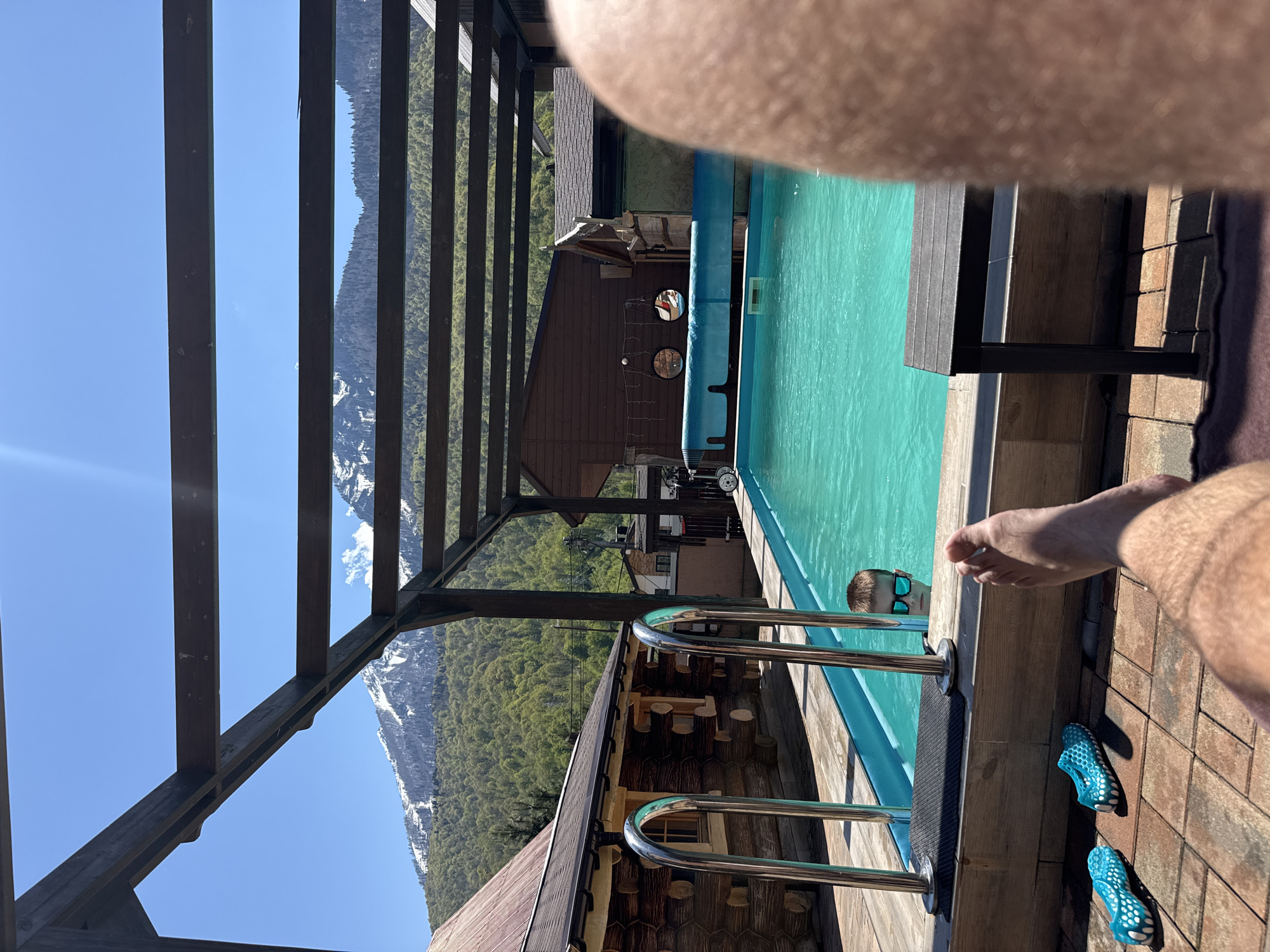 The photograph depicts an outdoor swimming pool area, possibly in Krasnaya Polyana, Russia, given the mountainous backdrop. The pool has a light blue hue, suggesting clean water. A set of stainless steel pool ladders are visible, extending into the water. A person is partially visible in the pool, wearing dark-rimmed sunglasses. A man is partially visible on the right, with his foot resting on the stone brick tiled deck. A pair of blue aqua shoes are sitting on the tiles next to him. The pool is situated next to a log cabin-style building with a brown roof. The scene is framed by a wooden pergola structure. In the background, a lush, green, mountainous landscape stretches towards the horizon, with snow-capped peaks partially visible. The sky above is clear and bright blue. The lighting indicates it is a sunny day.