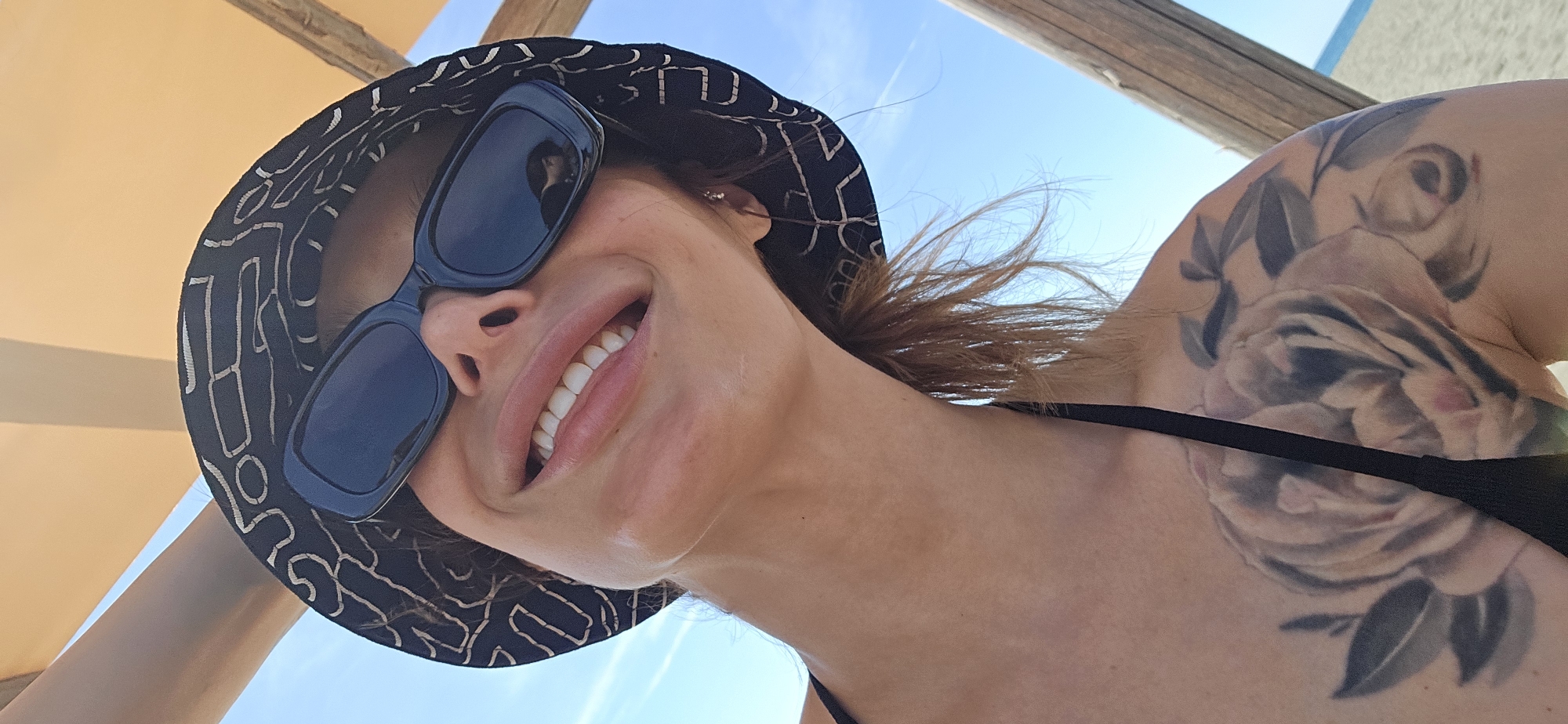 The image shows a woman, likely at a beach or similar sunny location, in the city of Gornji Štoj - Shtoji i Epërm, Montenegro. She is wearing a black bucket hat with a light-colored geometric design and dark sunglasses. Her visible skin is lightly tanned. There is a large floral tattoo on her upper right shoulder. She is smiling, revealing white teeth. The background includes a clear blue sky and a beige-colored beach shade structure. The shot is taken from a low angle, looking up at her face.