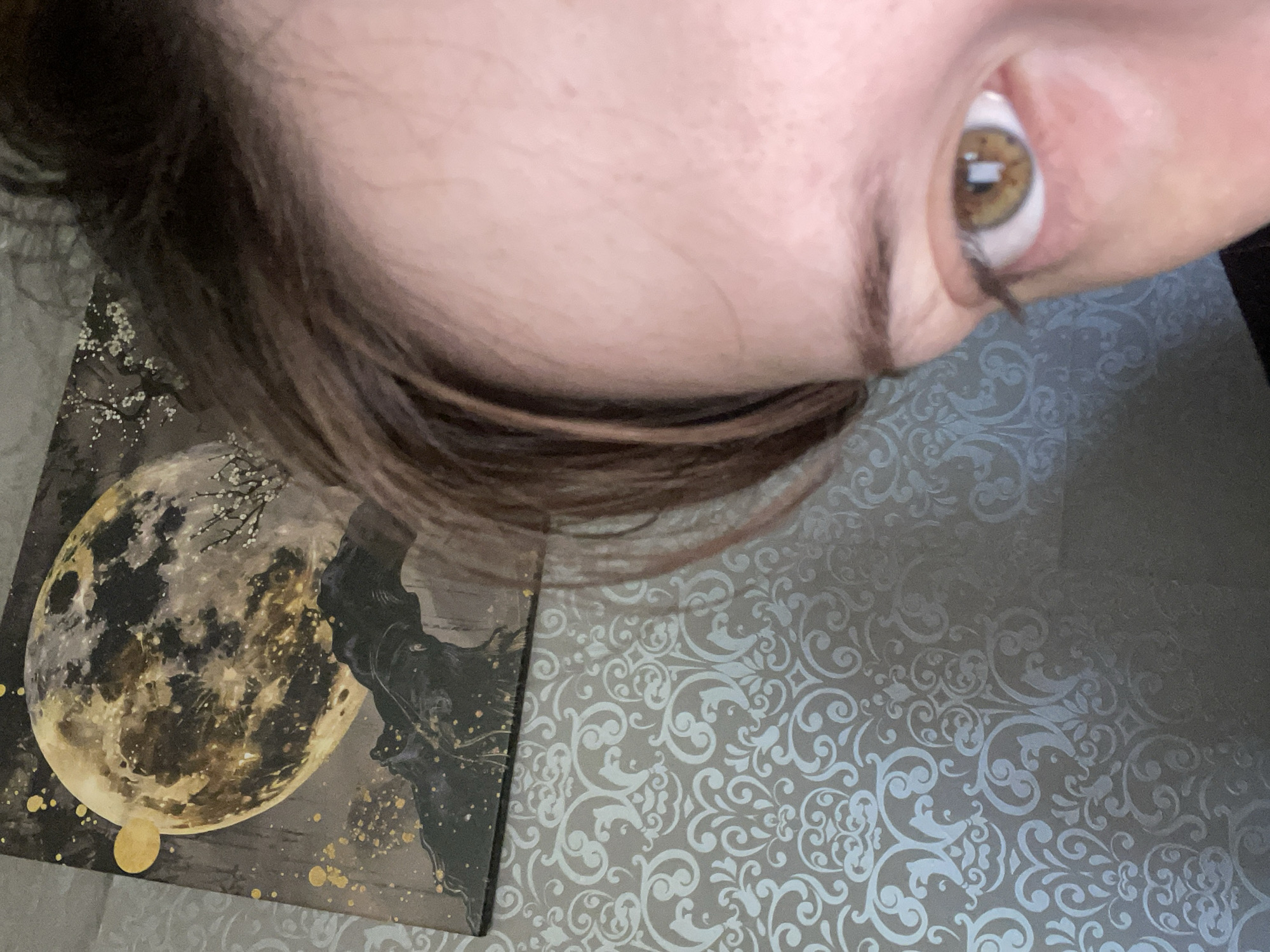 The image shows a close-up of a person's face, specifically focusing on their eye and forehead. The background consists of wallpaper with a decorative scroll pattern and a painting of a full moon with dark silhouetted mountains and a small tree with white blossoms. The person's hair is brown and falls across their forehead. The eye is light brown with visible reflections in the pupil. The lighting suggests it is an indoor setting. The image does not contain any information about the city of Линёвка, Russia.