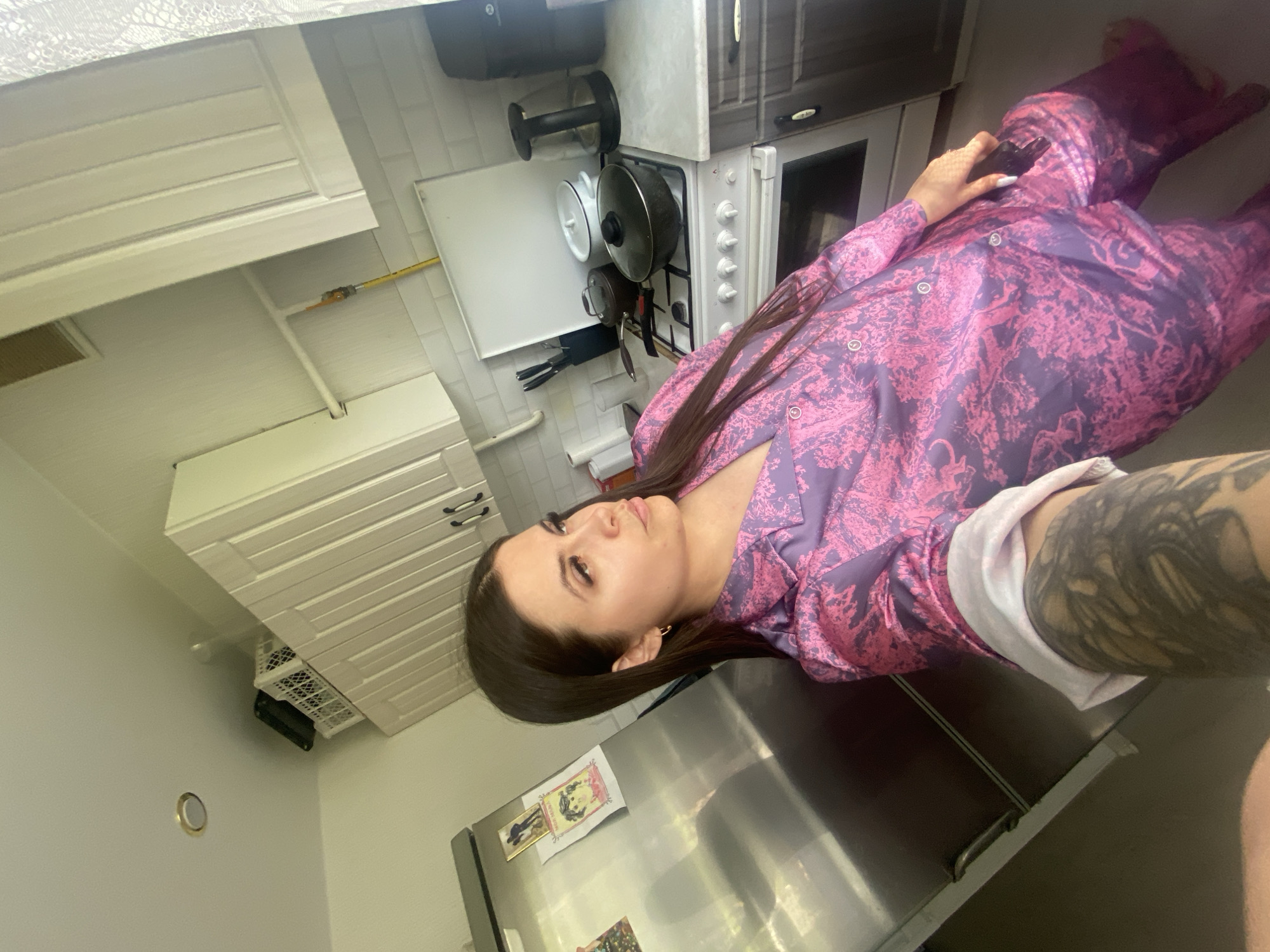 A woman is in a kitchen in Chaikovsky, Russia. She is wearing a patterned purple and pink pajama set. She is leaning against a stainless steel refrigerator. The kitchen has white cabinets and a white stove. There are pots and pans hanging above the stove. There are magnets and papers on the refrigerator.