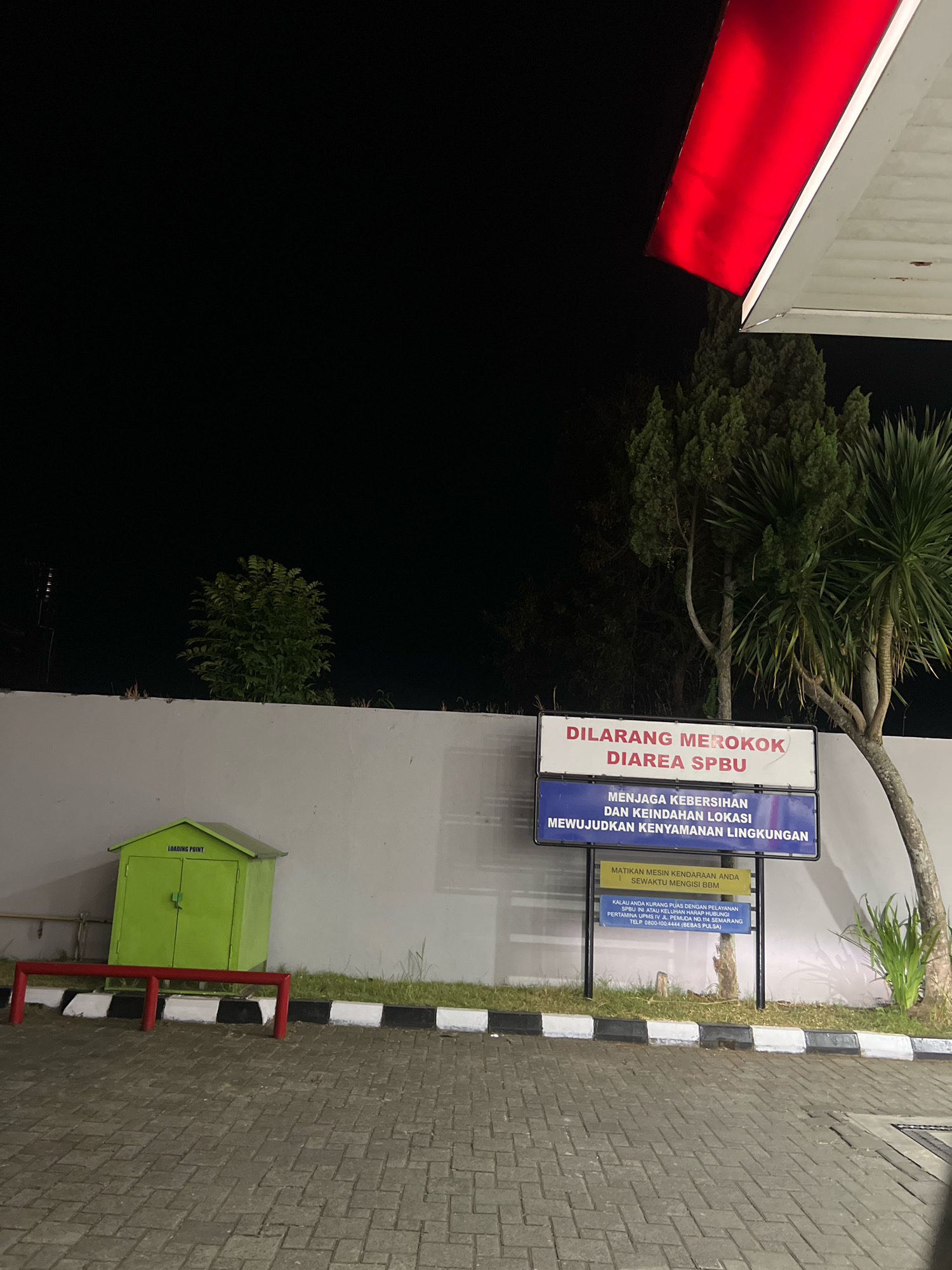 The image shows a night scene at what appears to be a gas station in Magelang, Indonesia. In the foreground, there is a paved area with interlocking brickwork. A red and white striped barrier runs along the edge of the paved area. To the left, there is a small, lime green utility box with "LOADING POINT" written on it. Behind the barrier and utility box is a white wall. Beyond the wall, trees are visible against a dark sky, with a bright red illuminated awning from a structure overhead.
There are two signs posted on a pole to the right of the green utility box. The top sign, in red lettering on a white background, reads "DILARANG MEROKOK DIAREA SPBU" (No Smoking in the SPBU Area). The second sign, in white lettering on a blue background, states "MENJAGA KEBERSIHAN DAN KEINDAHAN LOKASI MEWUJUDKAN KENYAMANAN LINGKUNGAN" (Maintaining the cleanliness and beauty of the location creates environmental comfort). A third, smaller yellow sign is below these, with instructions for customers.
There are no people visible in the image. The environment is dark, suggesting it is nighttime, and illuminated by artificial lights. The overall impression is that of a clean and orderly gas station area.
