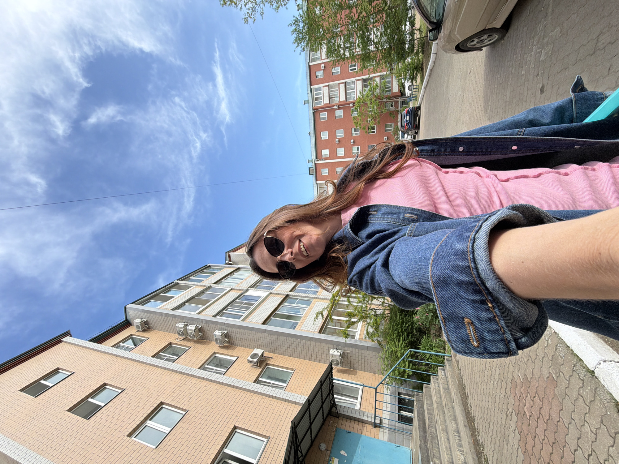 A woman with brown hair is taking a selfie outdoors. She is wearing sunglasses, a pink shirt, and a denim jacket. Behind her are two multi-story brick buildings, one yellow and one red. There is a blue sky with some clouds visible in the background. A sidewalk and some stairs are in the foreground. A car is partially visible on the right side of the image. The location is in the city of Birobidzhan, Russia.