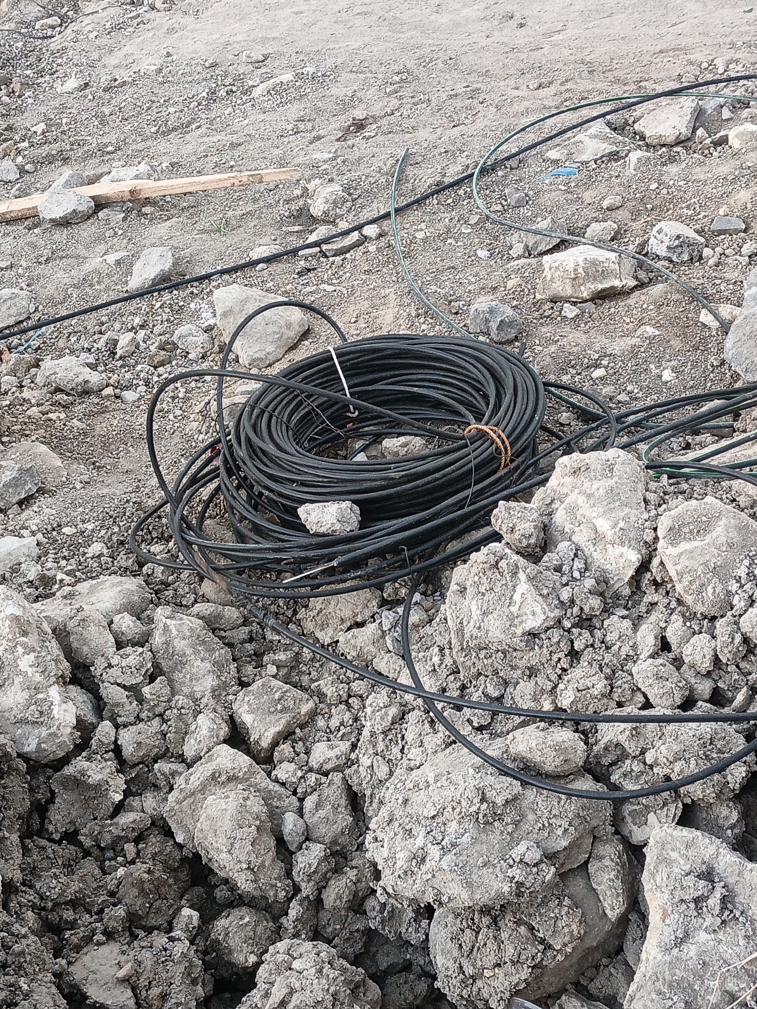 The image depicts a construction site in Planggu, Indonesia, with a focus on a coiled bundle of black cables lying on the ground amidst rocks and dirt. The cables appear to be thick and possibly electrical or telecommunication wiring, with some individual strands also visible extending outwards from the main coil and disappearing into the surrounding debris. The environment is characterized by loose soil, gravel, and a variety of greyish rocks, suggesting recent excavation or groundwork. A small piece of light-colored wooden debris is present in the upper left quadrant of the image. There are no people or animals visible in the scene. The overall impression is one of an active or recently disturbed construction area.