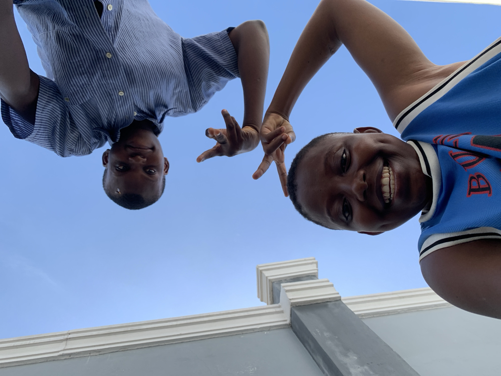This low-angle photograph, taken in Ibasa, Nigeria, features two young individuals looking down into the camera against a bright, clear blue sky. The person on the left wears a blue and white striped short-sleeved shirt and has a gentle smile. The individual on the right, wearing a blue top with white and black trim, displays a wide, joyful smile, showing their teeth. Red lettering, partially visible as a "B" or "R", is present on the right person's shirt. Both subjects extend their hands towards the camera, making a two-finger gesture resembling a peace sign. Below them, a light gray wall with white architectural coping and a matching column is visible, suggesting an outdoor setting. The absence of clouds indicates clear weather during daytime. The expressions of both individuals convey happiness and playfulness.