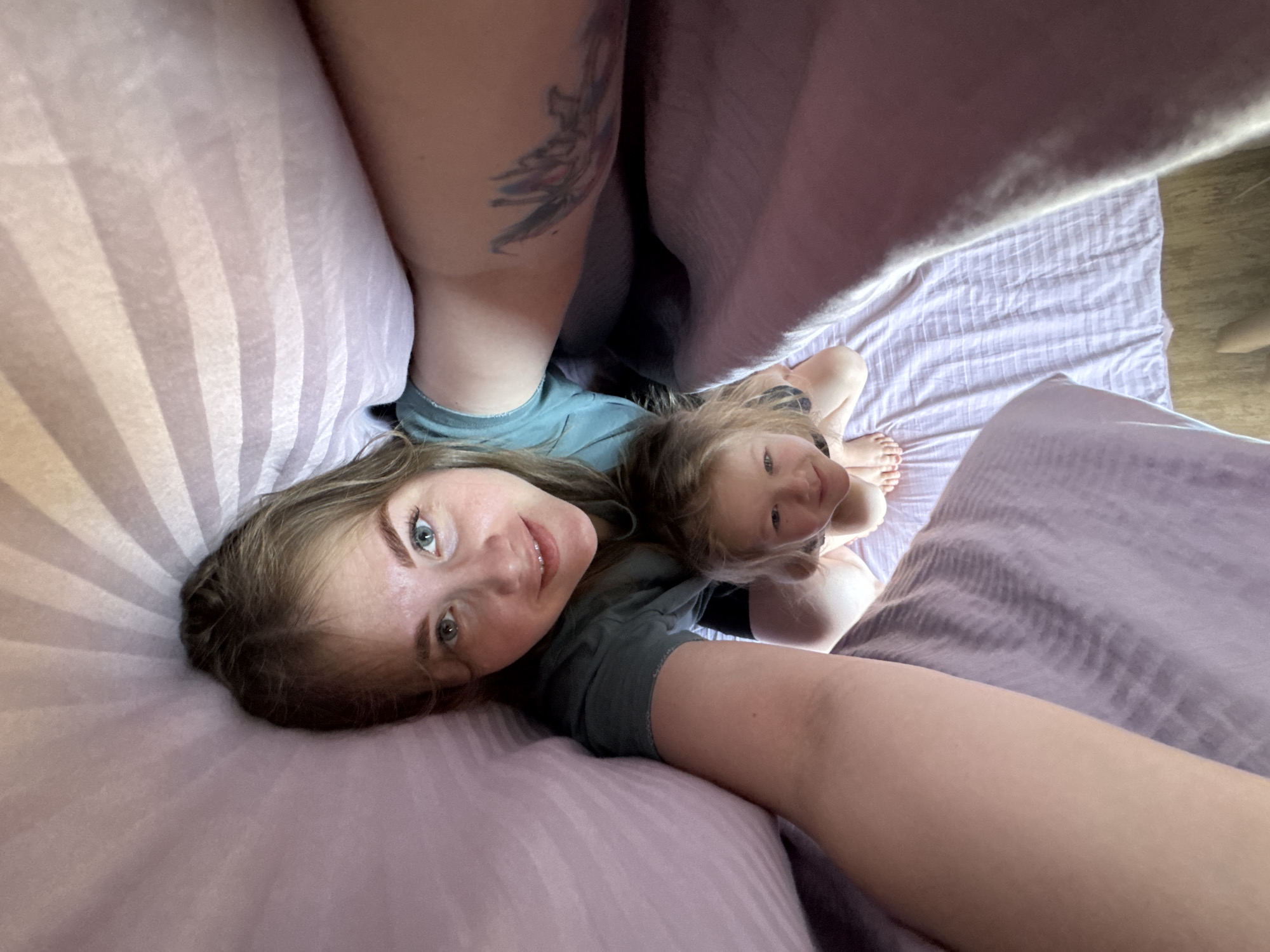 Two individuals are visible in the image. An adult female with fair skin, light brown hair, and blue eyes is positioned close to the camera, partially lying on a light pink, striped pillow. A smaller child, presumably female, with light hair, is positioned behind and slightly above the adult, also looking at the camera. Both are wearing what appear to be t-shirts; the adult's is a dark green.
The scene is likely indoors on a bed, indicated by the pink and light purple striped bedding. The perspective is from above, as if someone is taking a selfie with a camera held at an angle looking down.
The image does not include enough information to confirm that the image was taken in the city of Ферма, Russia.