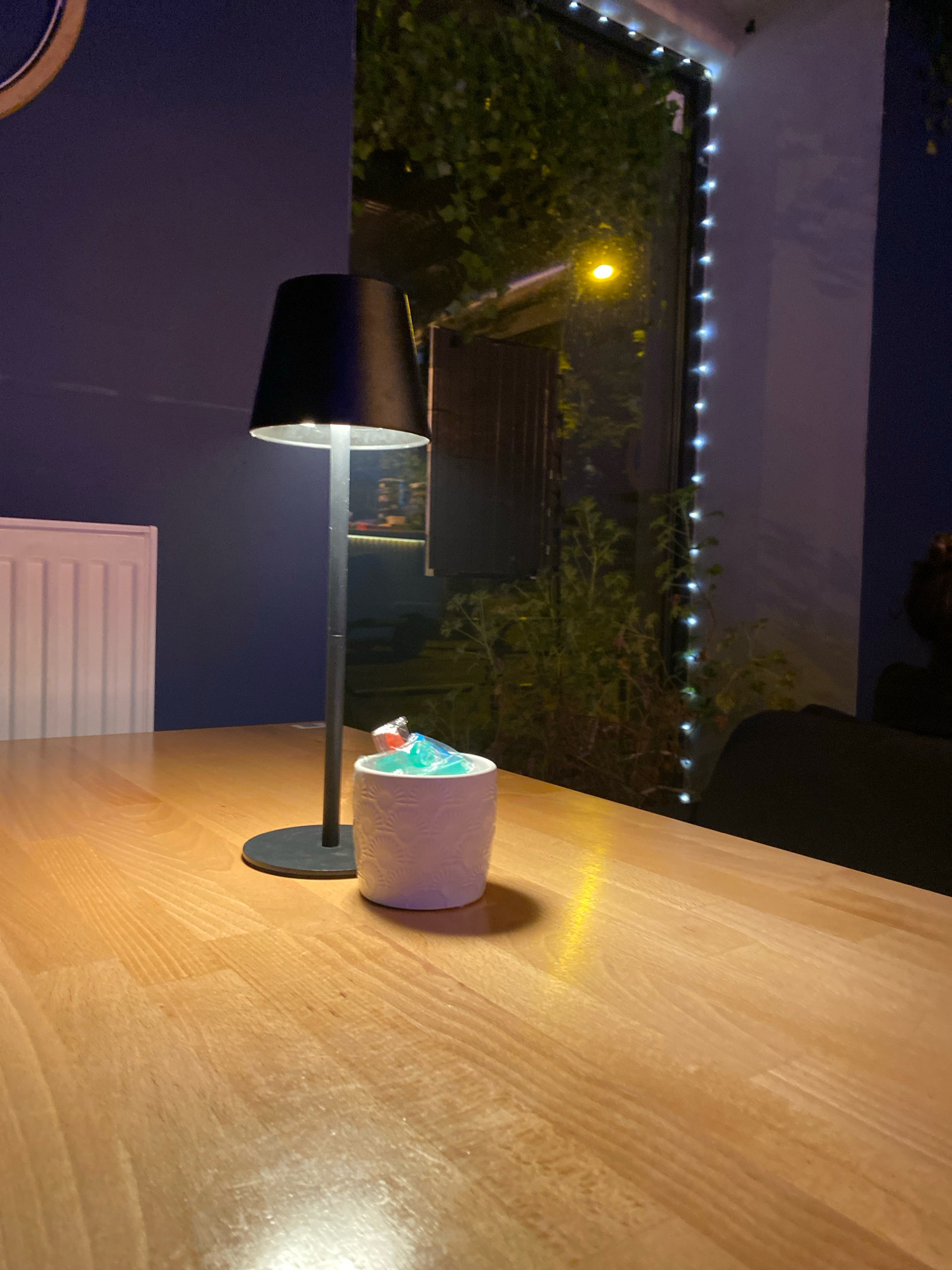 The image depicts a dimly lit interior scene, likely a restaurant or cafe, with a wooden table in the foreground. On the table, a modern black lamp with a conical shade is illuminated. Beside it sits a small, white textured container filled with brightly colored candies, some appearing to be wrapped in cellophane. In the background, a window reveals an outdoor area with dense greenery and faint streetlights or decorative lights. The wall behind the table is a deep blue. To the left, a white ribbed radiator is partially visible. The overall ambiance suggests an evening setting. This scene could be from a restaurant in Gliwice, Poland, given the context of the request.