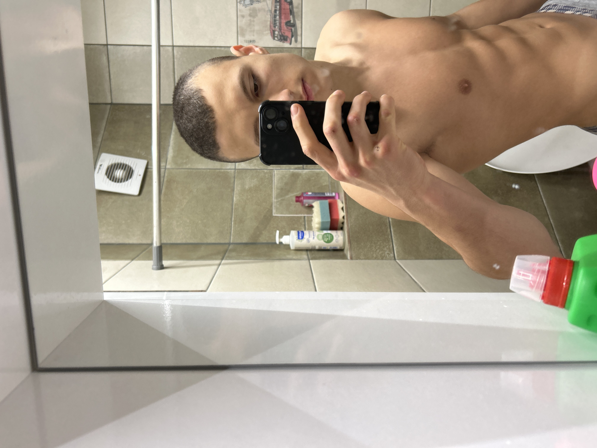 A shirtless male takes a selfie in a bathroom mirror. He is holding a black smartphone. His abs are visible. The bathroom has tiled walls and floors in a light beige tone. Behind him, cleaning supplies and a white ventilation fan can be seen on the floor. There is also a tall, thin pole, possibly a shower support. The location, Burgas, Bulgaria, cannot be confirmed solely based on the image.