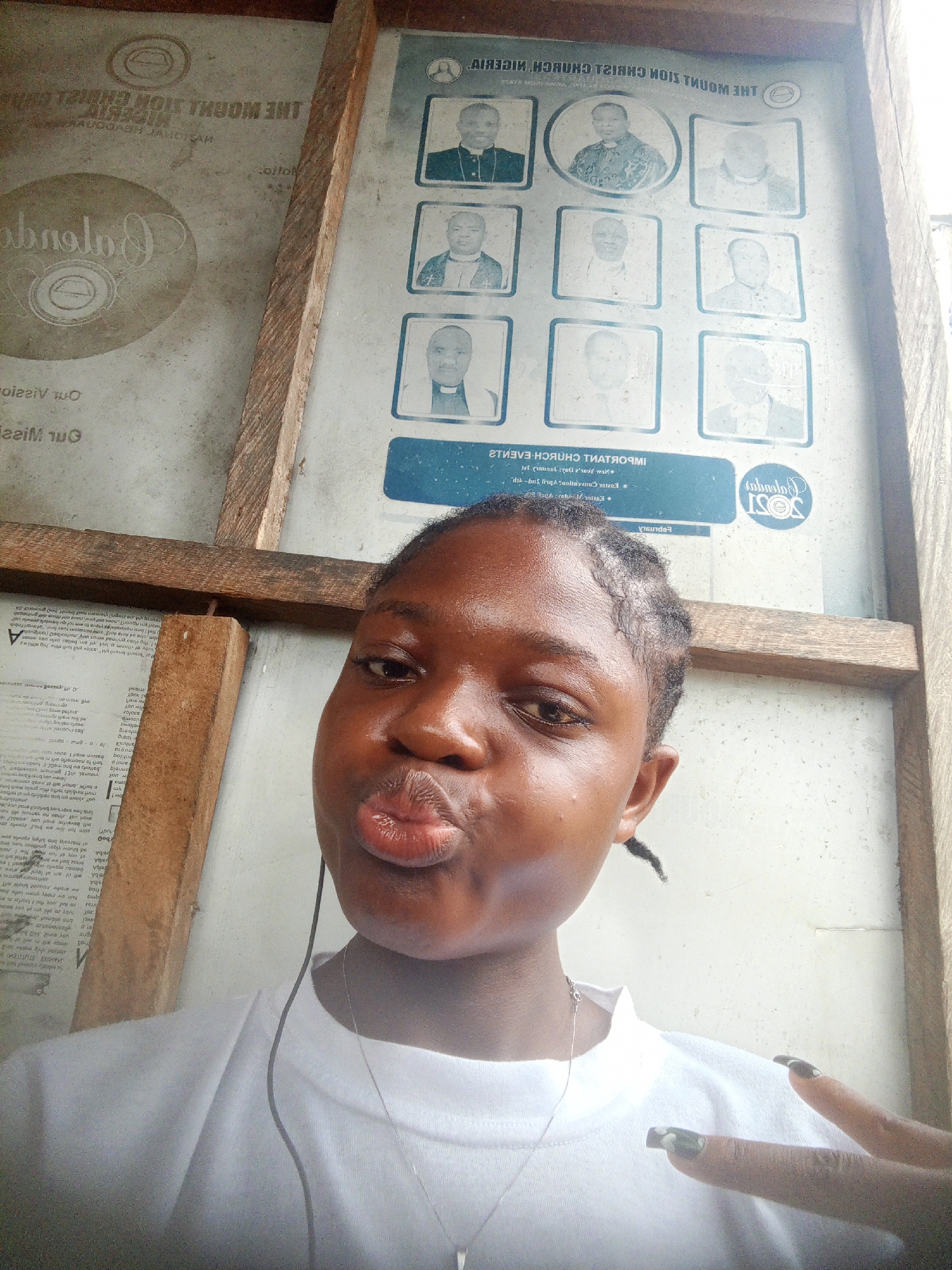 A young woman with braided hair, wearing a white shirt and a silver necklace, is taking a selfie. Her lips are pursed in a kissing gesture, and she holds up two fingers in a "peace" sign. She stands in front of a wooden frame and a display board.
The display board contains two visible sections. On the right, framed portraits are arranged in a 3x3 grid. Above the grid, the text "THE MOUNT ZION CHRIST CHURCH NIGERIA" is visible. Below the grid, a blue banner displays the words "IMPORTANT CHURCH EVENTS". A logo is also present, featuring "2021".
To the left, a separate display area features a large, circular logo. The surrounding text indicates the location is Mbribit, Nigeria. There's also printed text underneath the logo.