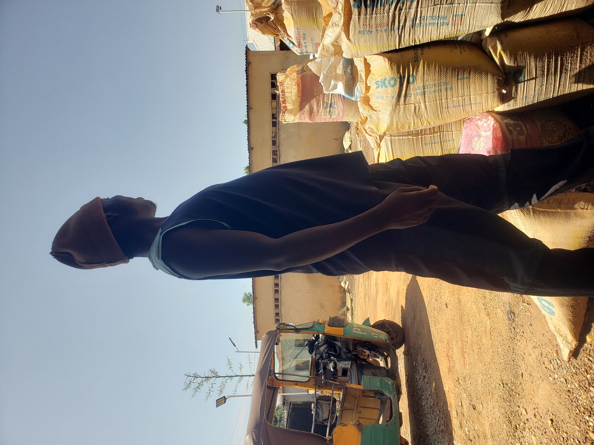 A young man wearing a brown beanie and a dark blue sleeveless shirt stands in an outdoor setting, looking to his right. He is partially obscured by shadows and is positioned in the foreground, facing away from the camera. Behind him, several large sacks are stacked high, appearing to contain cement or similar materials. The sacks are predominantly light brown and yellow with some text visible, including "SOKOYO CEMENT" and "AL-AZIZ CEMENT".
To the left of the man and the sacks is a three-wheeled vehicle, possibly an auto-rickshaw, painted in green and yellow. The vehicle has a covered top and appears to be parked on a dirt and gravel surface. In the background, there is a long, light-colored building with a row of small windows or vents. The sky is clear and bright blue, suggesting a sunny day. The overall environment appears to be a storage or market area.
Based on the presence of specific cement brands like "SOKOYO CEMENT" and "AL-AZIZ CEMENT", which are known to be produced and distributed in Nigeria, and considering the general visual cues of the environment (e.g., architecture, vehicle type), this scene is consistent with a location in Jos, Nigeria. The scene depicts a man possibly involved in loading or unloading goods, given the context of the sacks and the vehicle.