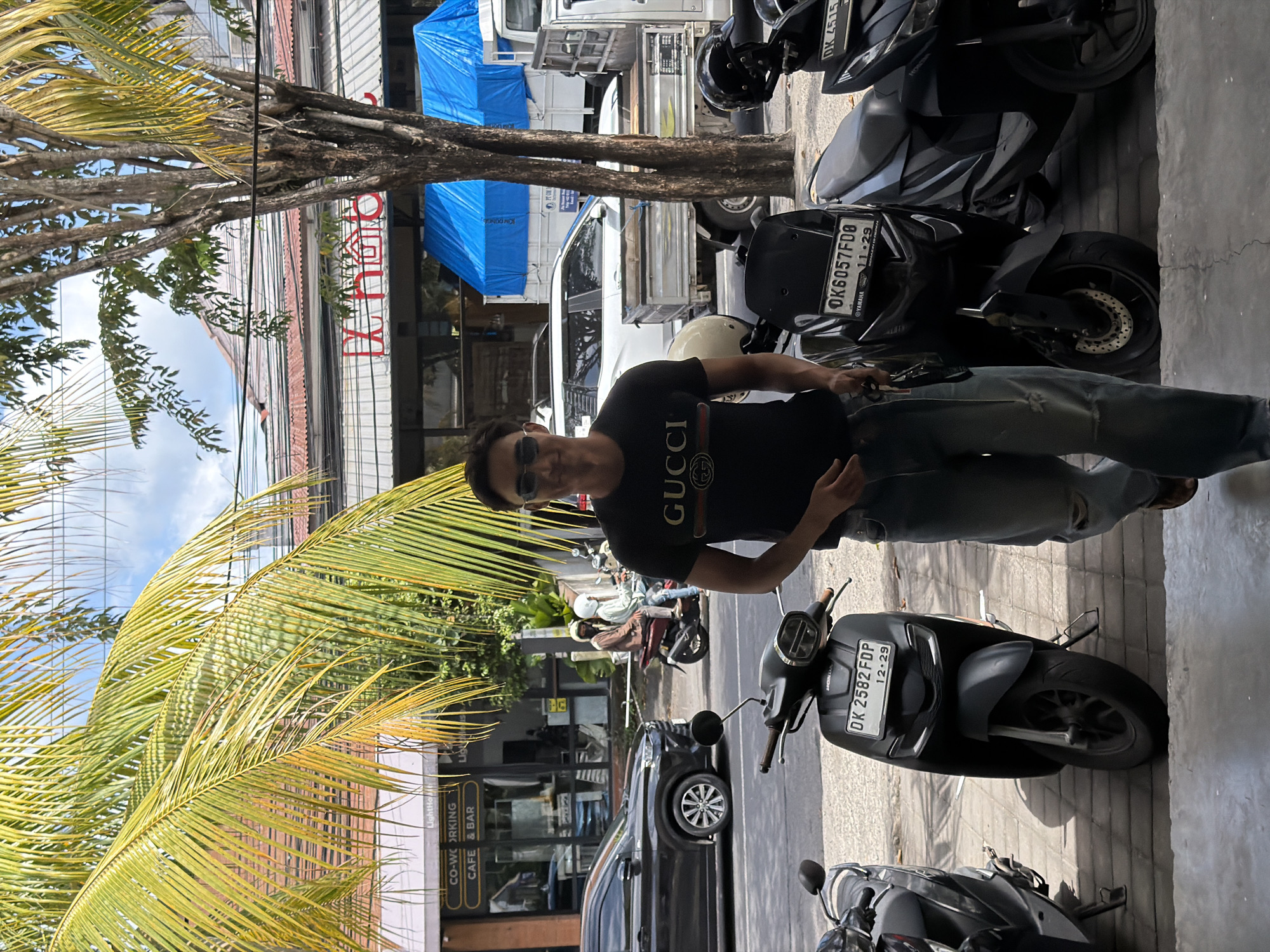 The image captures a scene in North Kuta, Indonesia, with a man posing on a street lined with parked motorbikes and buildings. The man is wearing a black Gucci t-shirt, sunglasses, and jeans. He stands beside a grey Honda motorbike with the license plate "DK 2582 FDP 12-29." Several other motorbikes are parked along the street, including one with the license plate "OK6057FD8 11-29" from Yamaha. In the background, a building labeled "LightHolt CO-WORKING CAFE & BAR" is visible. A palm tree is on the left side of the image, adding to the tropical atmosphere. Cars and other motorbikes are also seen in the background, indicating a busy street. A building with blue tarps covering a portion of it can be seen towards the upper part of the image.