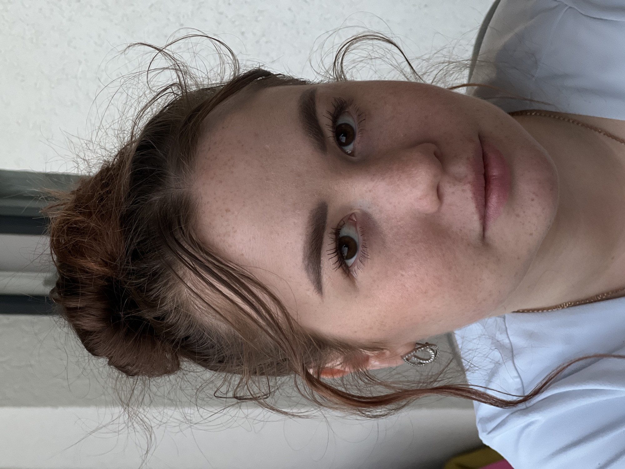 A close-up, head-and-shoulders portrait captures a young woman with auburn hair pulled into a messy bun. She has fair skin with freckles, brown eyes, and light brown eyebrows. She is wearing a light blue t-shirt and a necklace with a gold chain. In her left ear, she has a silver teardrop-shaped earring with small stones. Her hair is windswept, with several strands falling around her face and ears. The background is a textured white wall, suggesting an outdoor setting or a room with a plain wall. There is no indication of the city of Florești, Romania, or any specific urban environment.