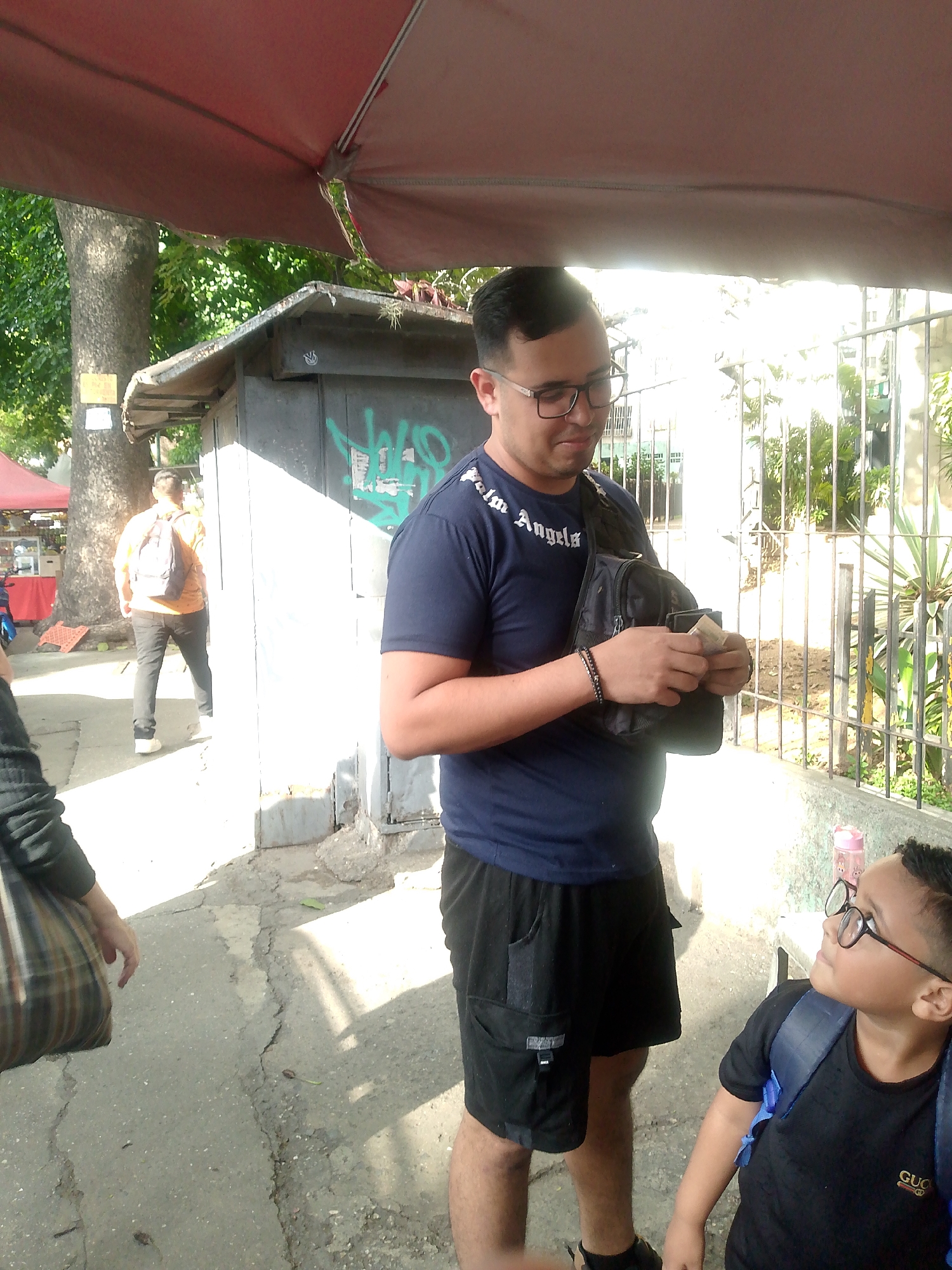 The photo shows two people, one adult and one child, standing in front of a weathered booth with graffiti on a city sidewalk in Caracas, Venezuela. The adult male, wearing glasses, a dark t-shirt with “Palm Angels” written on it, black shorts, and a shoulder bag, is counting money. The child, wearing glasses, a black t-shirt, and a backpack, is looking up at the adult. The scene is lit by natural sunlight, with a red awning partially visible overhead. A metal fence is present to the right of the subjects. In the background, a person walks away from the camera on the sidewalk near a market stall set up under trees.