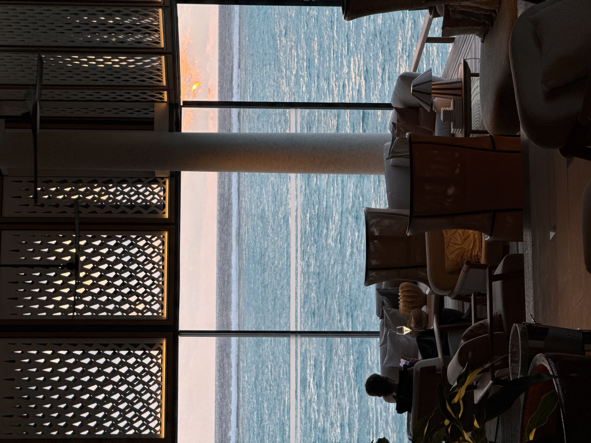 A person is seated on a sofa, looking at their phone, with a glass of white wine on a small table beside them. The scene is set indoors, with large windows offering a view of the ocean and a sunset. Decorative panels with a diamond pattern are visible above the windows, and a ceiling fan is suspended from the ceiling. The room is furnished with sofas and chairs, a small table with a lamp, and decorative items. The overall atmosphere is serene and relaxing. The location is identified as Unknown, Maldives.