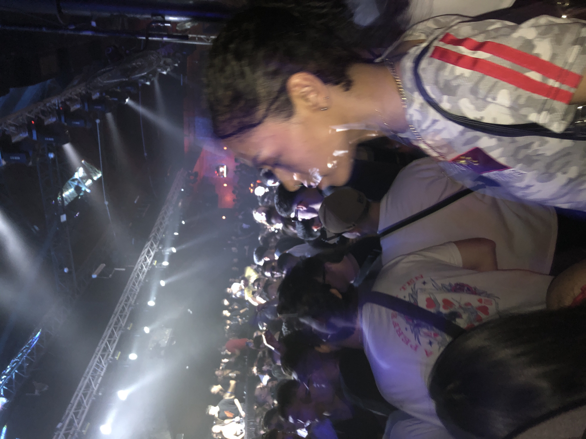 The image shows a crowded event, possibly a concert, with a large crowd of people. The lighting is dim with spotlights illuminating the crowd. A metal structure is visible in the background, likely part of the stage rigging. A person in a camouflage shirt with red stripes is in the foreground. The event appears to be taking place indoors. The exact location in Buenos Aires, Argentina, cannot be determined from the image.