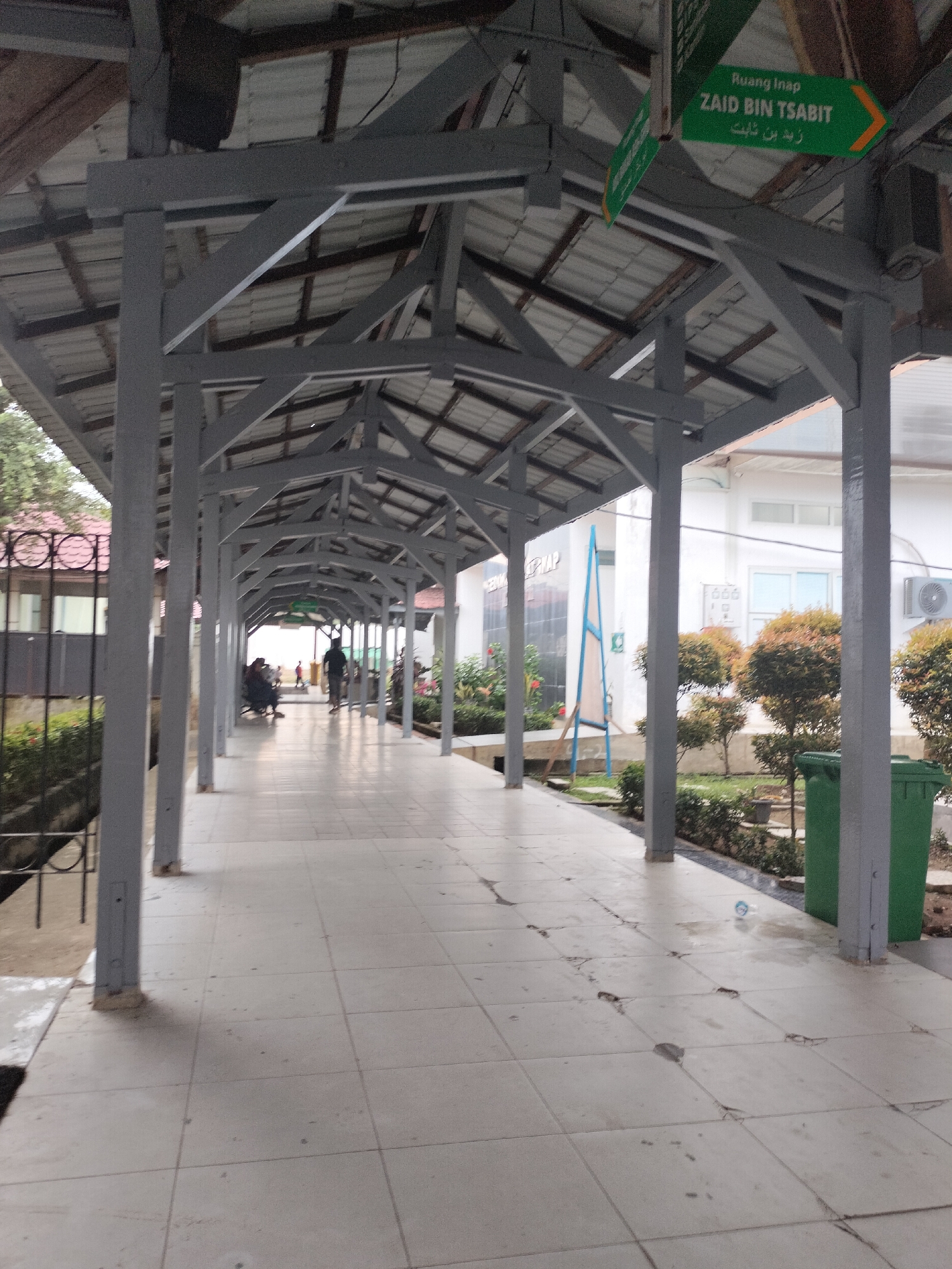 The image depicts an outdoor covered walkway at the Langsa City General Hospital in Langsa, Indonesia. The walkway is constructed with a gray metal frame and a corrugated metal roof, supported by numerous gray pillars. The floor is tiled with off-white square tiles.
In the foreground, the walkway stretches into the distance, with a few people visible in the middle ground. On the left side, near the entrance of the walkway, several individuals are seated on a bench. Further down the walkway, a person is walking away from the camera.
The walkway is bordered by greenery on the right side, including neatly trimmed bushes and some flowering plants. A bright green trash bin is positioned near the end of the walkway on the right. On the left side of the walkway, there is a fence and some trees.
A green directional sign is visible hanging from the roof structure in the distance. The sign has text in Indonesian and Arabic, with an arrow pointing to the right. The text appears to read "Ruang Inap ZAID BIN TSABIT".
The overall environment suggests a hospital setting, with the covered walkway providing shelter and a path between buildings. The lighting indicates it is daytime, though the sky is not visible. The condition of the tiles on the floor shows some wear and tear.