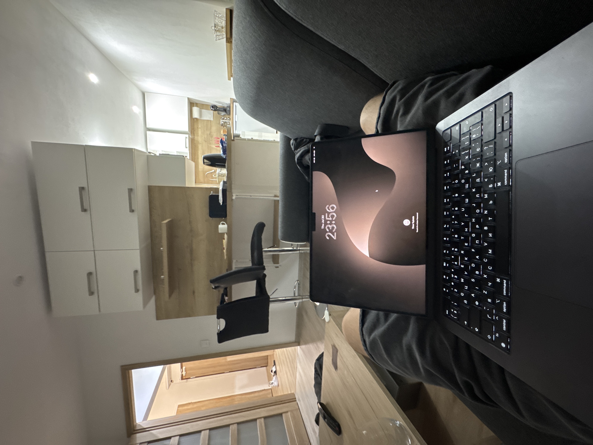 The image shows a person sitting on a dark gray sofa with a laptop open on their lap. The laptop screen displays the time as 23:56 and the date as Thu Jul 30. The background suggests an indoor living space with a kitchen area visible, featuring white cabinets, a wooden countertop, and kitchen appliances. To the left, a doorway leads to another room with a table and chairs. The overall environment appears to be a modern apartment or house. The location is not specified within the image itself.