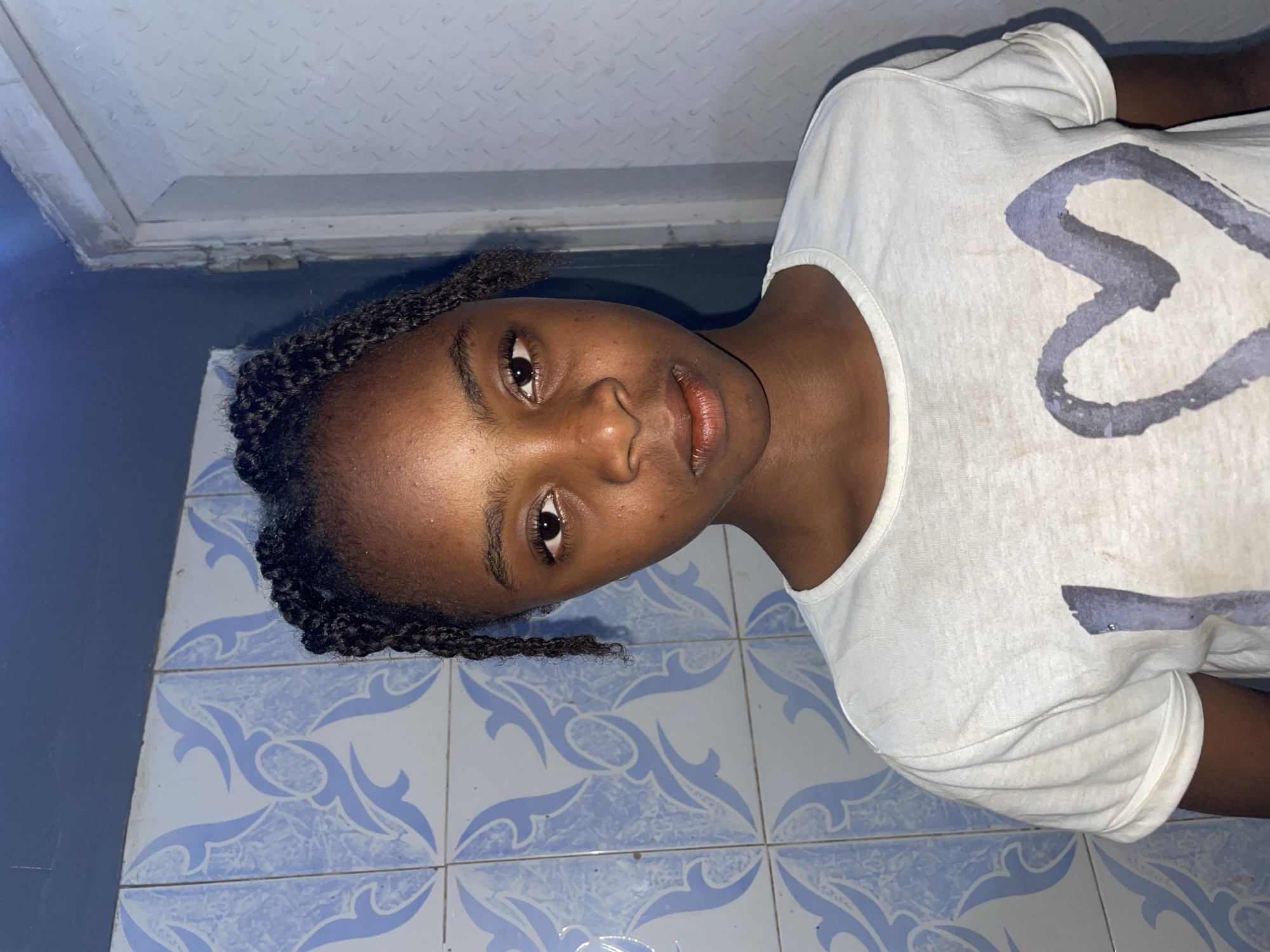 A young, dark-skinned person lies on their side, facing the camera. They have dark, braided hair, and are wearing a white t-shirt with a faded grey design on the front. The background consists of blue-and-white patterned tiles, a dark blue wall, and a white ceiling with a textured or slightly damaged surface. There is no information available to confirm the location in Jos, Nigeria.