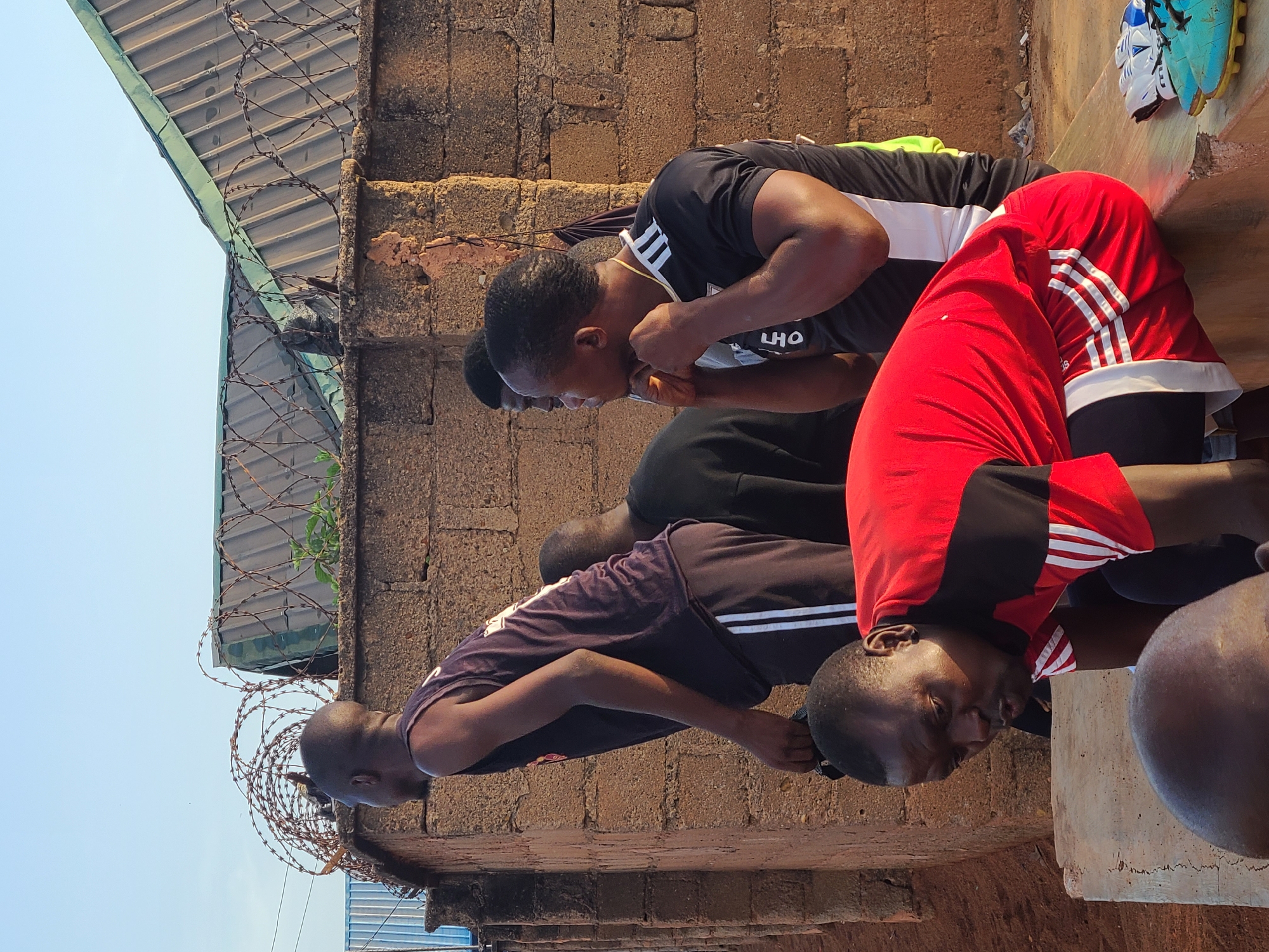 A group of men stand in a row, bending at the waist. They are in front of a brick wall topped with barbed wire. A corrugated metal roof is visible above the wall. The men are wearing various athletic shirts, including red, black, and purple with stripes. On a ledge next to them sit a pair of soccer cleats. The location is the city of Unknown, Unknown.