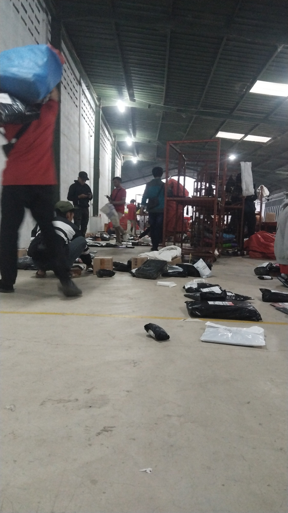 The image depicts a scene inside a warehouse or distribution center, likely located in Tarajusari, Indonesia. Several individuals, appearing to be workers, are engaged in various activities related to package handling. In the foreground, a person is crouched down, seemingly sorting or inspecting packages on the concrete floor. Numerous packages, mostly black and white, are scattered across the floor, some appearing to be sealed in plastic, while others are in cardboard boxes.

Several other individuals are visible throughout the space. One person in the left foreground is carrying a large blue bag over their shoulder, along with other packages. Further back, individuals are standing and interacting, with one person appearing to hold up a package. Racks and shelves are present in the background, indicating organized storage. The environment appears to be dimly lit with overhead lighting, and the ceiling has a corrugated metal structure. The overall impression is one of an active logistics or fulfillment operation.