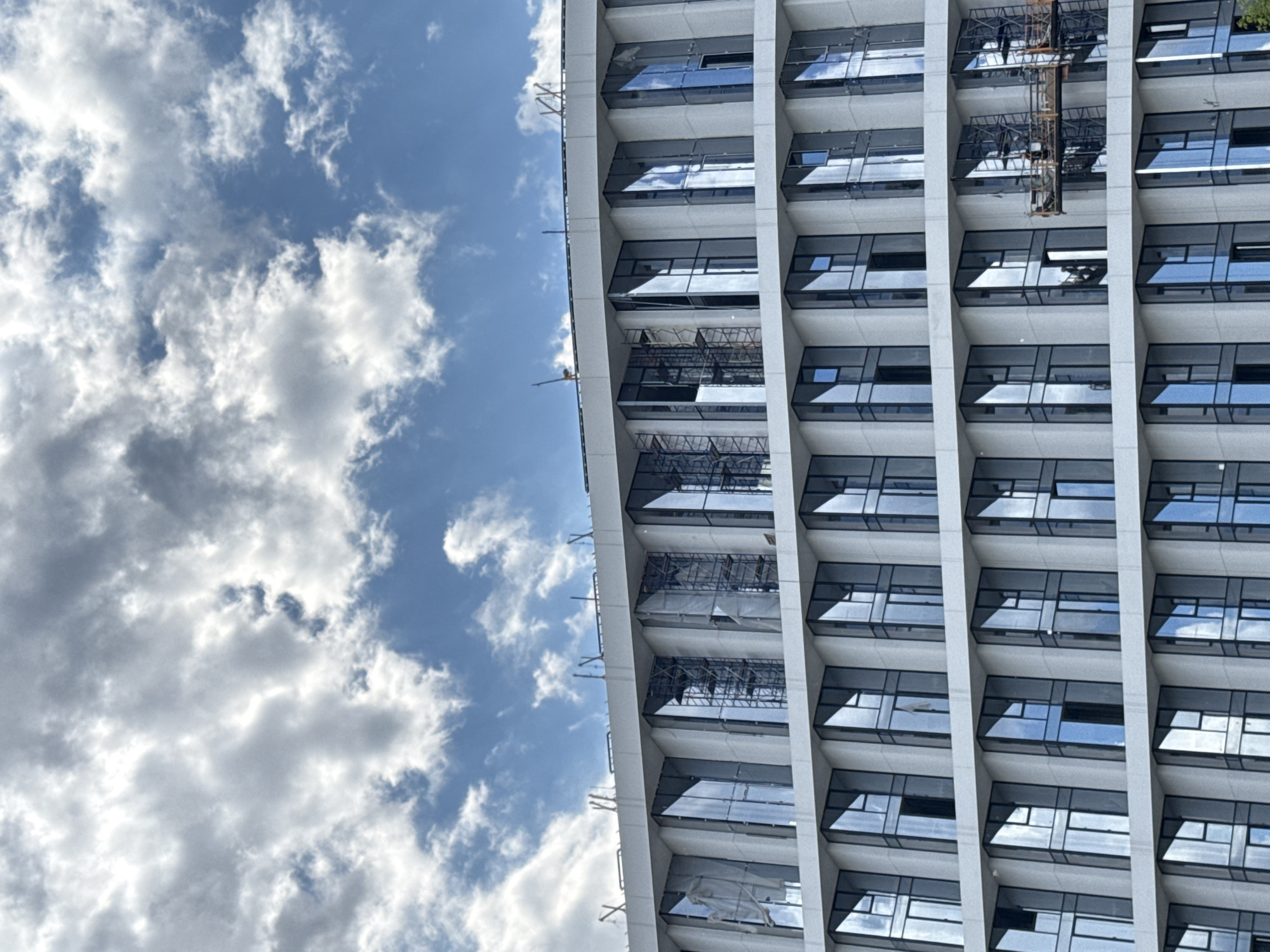 This image captures a modern, high-rise building against a bright blue sky filled with scattered cumulus clouds. The building, which occupies the right side of the frame, features a grid-like facade composed of reflective dark-tinted windows set within light-colored vertical and horizontal structural elements. The windows distinctly reflect the sky and clouds.
Evidence of ongoing work or construction is visible on multiple floors. Scaffolding and temporary platforms are attached to the building's exterior, particularly on the left side of its visible portion. One section clearly shows a suspended platform with intricate metalwork. Additionally, white protective coverings or tarps are present on some levels. A small yellow crane arm extends from the upper left edge of the building.
The sky on the left side of the frame is a clear blue, with fluffy white and grey clouds of varying densities, indicating a sunny day with some cloud cover. There are no people visible in the image, and no text is discernible. The scene depicts a contemporary architectural structure, likely under construction or undergoing maintenance, in an urban setting identified as Moscow, Russia.