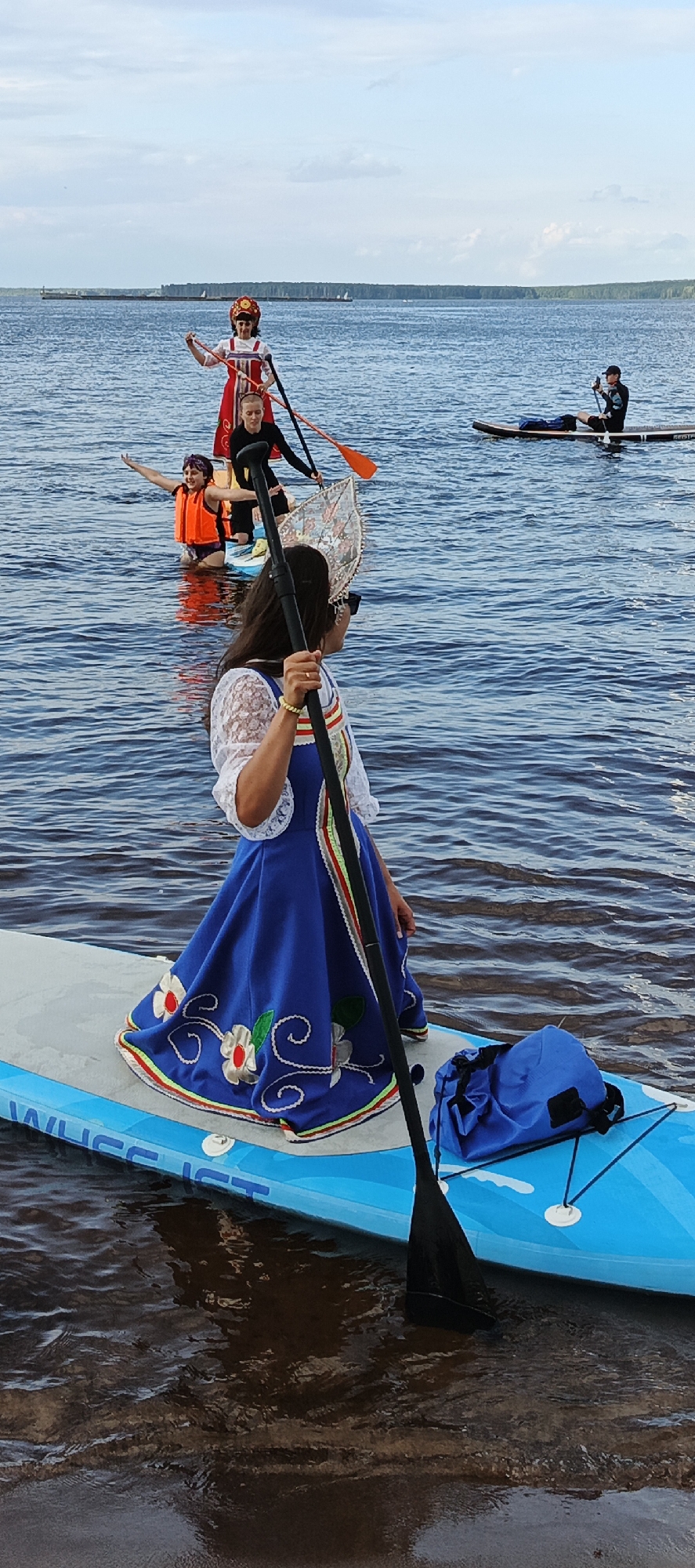 In a lake near Chaikovsky, Russia, several people are paddleboarding. The foreground features a woman in a traditional blue and white folk dress adorned with floral embroidery and a decorative headpiece. She holds a paddle, her gaze directed to the side. Behind her, other paddleboarders are visible. One individual in an orange life vest is waving, while another person in a red and white outfit stands on a paddleboard. Further in the background, a person is seated on a paddleboard. The water is calm and reflects the overcast sky, suggesting it is daytime. The overall mood appears to be festive or recreational. The paddleboard in the foreground has "VAIUS ICT" visible on its side.