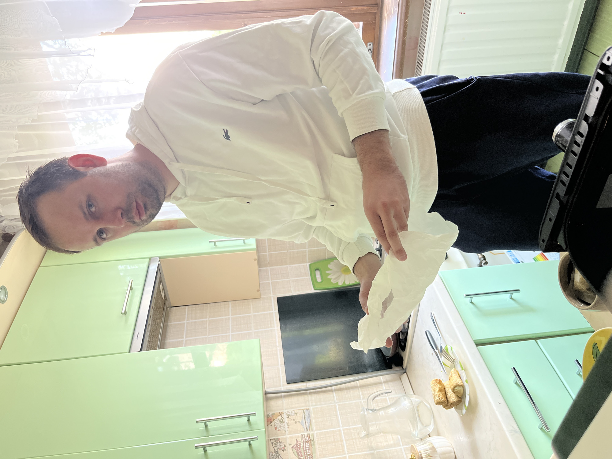 The image shows a man standing in a kitchen. He is wearing a white hooded sweatshirt with a logo on the chest and dark-colored pants. He is holding a crumpled white piece of paper. The kitchen has light green cabinets and drawers with silver handles, a black stovetop, and a beige countertop. A glass pitcher and a small plate with pastries are on the counter. The location is likely a residential kitchen, but there are no specific details that confirm that it is in the city of Rusanovo, Russia. The environment appears to be well-lit, likely by a window covered with a white curtain.