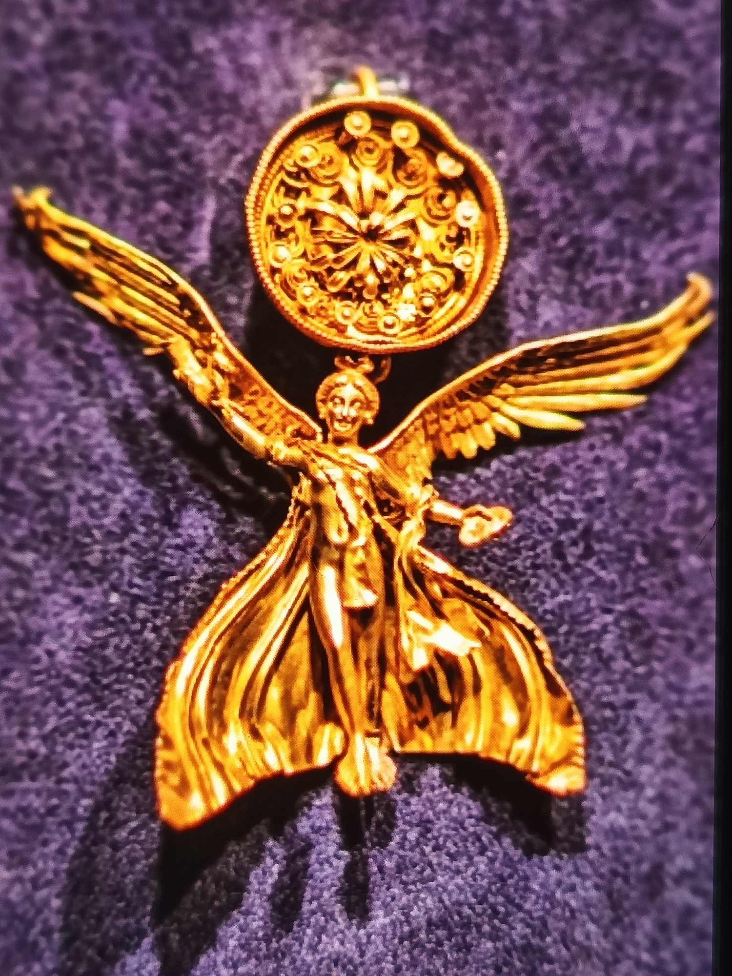 A close-up, top-down, eye-level shot shows a gold pendant in the shape of a winged figure, possibly the goddess Nike, with its wings spread wide and its drapery flowing. The pendant is resting on a textured, deep purple surface, which appears to be fabric, possibly velvet. Above the winged figure is a circular medallion with intricate gold detailing, resembling a starburst pattern. The figure itself is slender and appears to be holding something small in its outstretched right hand. The lighting creates strong highlights and shadows on the metallic surface, emphasizing the texture and detail of the pendant. This artifact is housed in the Archaeological Museum in Varna, Bulgaria.