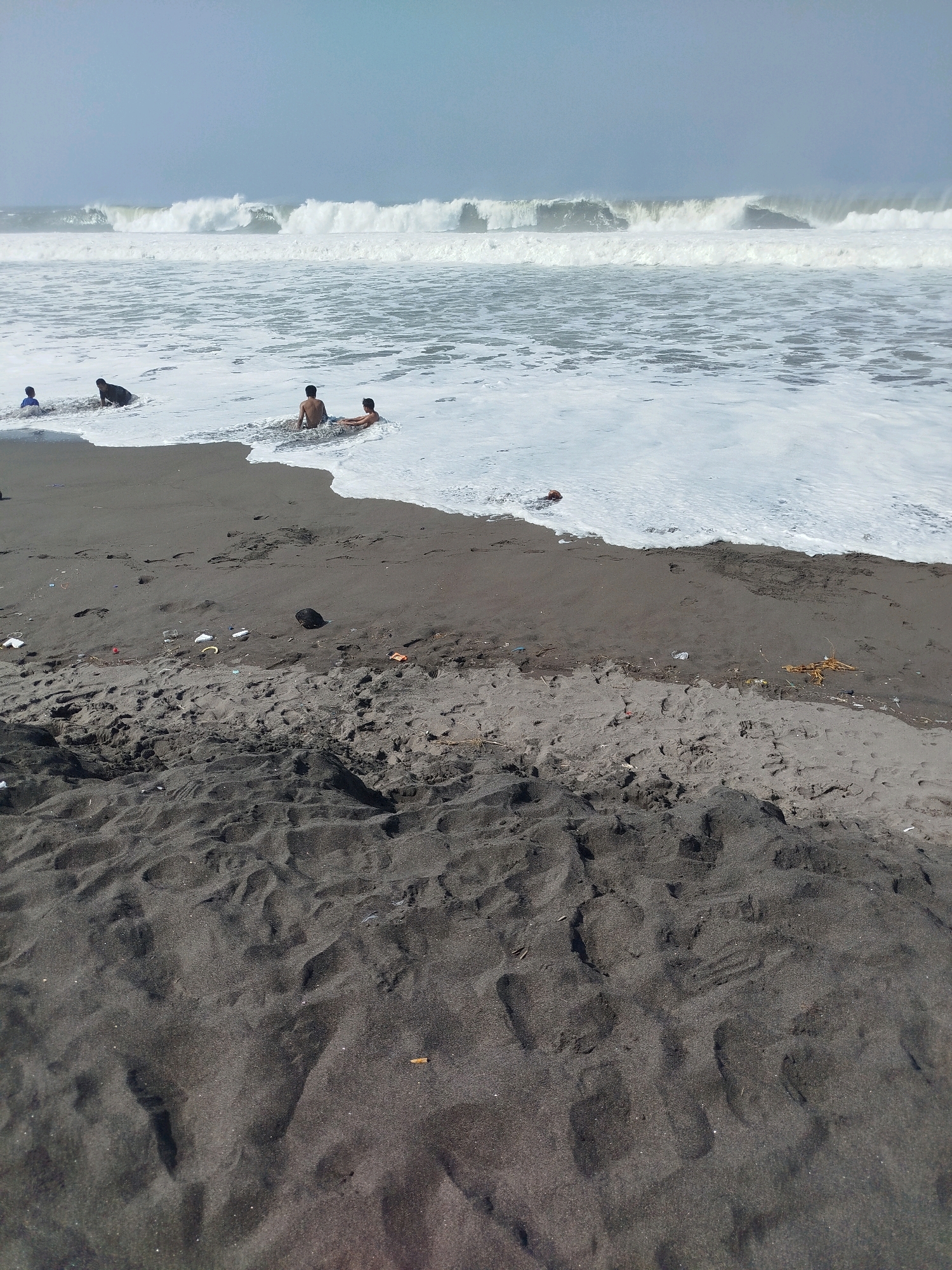 A wide shot captures a beach with dark sand leading to the ocean. Several people are in the shallow water near the shore, some appearing to be lying down or sitting. Large waves crash in the distance under a hazy sky. Debris, including plastic and organic matter, is scattered on the sand closer to the foreground. The overall scene suggests a natural beach environment impacted by pollution. This location is identified as Kenoyajaya, Indonesia.