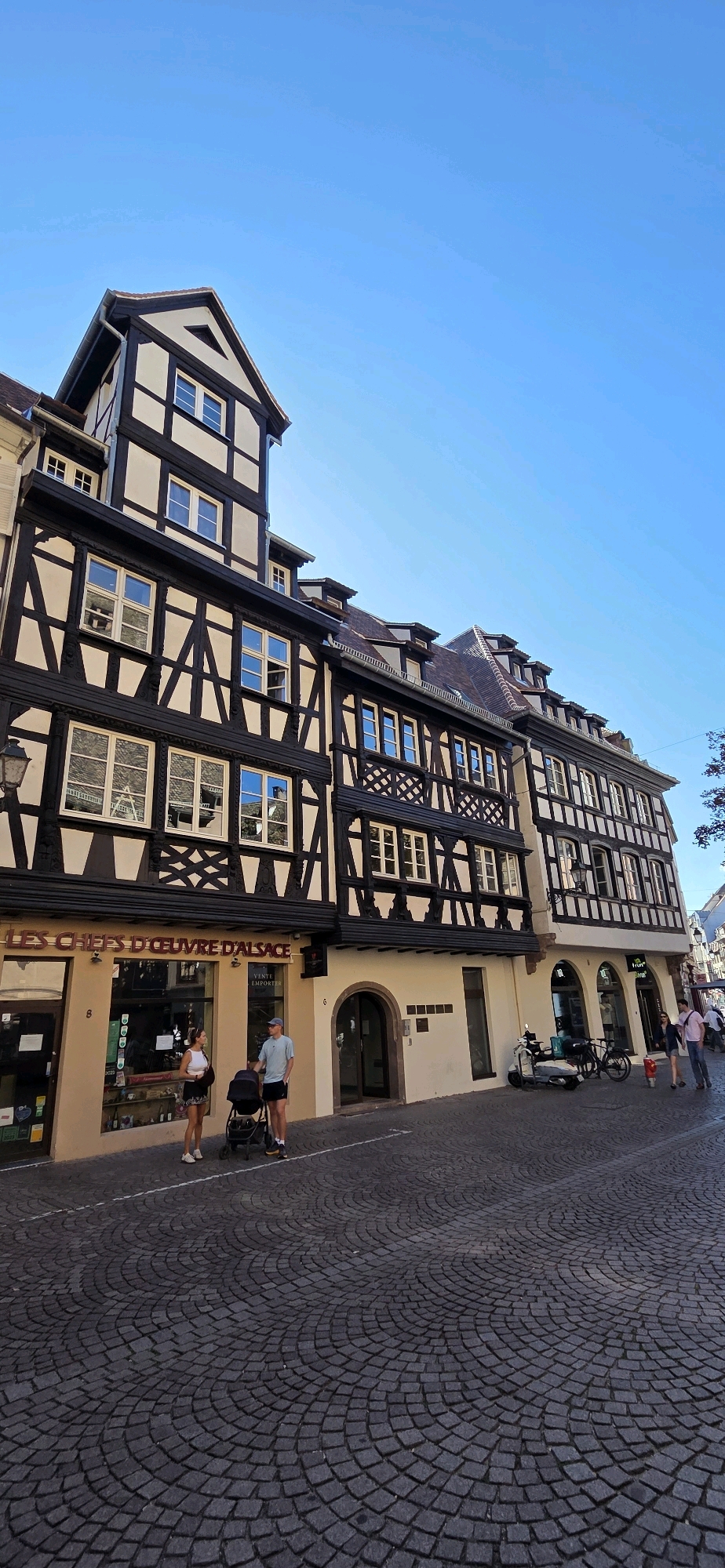 The image depicts a street scene in Strasbourg, France, with a focus on the distinctive half-timbered architecture characteristic of the region. The buildings feature dark wooden beams against a lighter facade, with numerous windows. The street is paved with dark cobblestones laid in a radial pattern.
In the foreground, a couple stands with a baby stroller. The woman is wearing a white top and a dark skirt, while the man is in a light blue t-shirt and dark shorts, wearing a baseball cap. They are standing in front of a shop with the text "LES CHEFS D'OEUVRE D'ALSACE" visible on its facade.
Further down the street, a few other people are walking, and a scooter and a bicycle are parked near an arched doorway. The sky is a clear, bright blue, suggesting good weather. The overall scene captures the charm of a European city street.