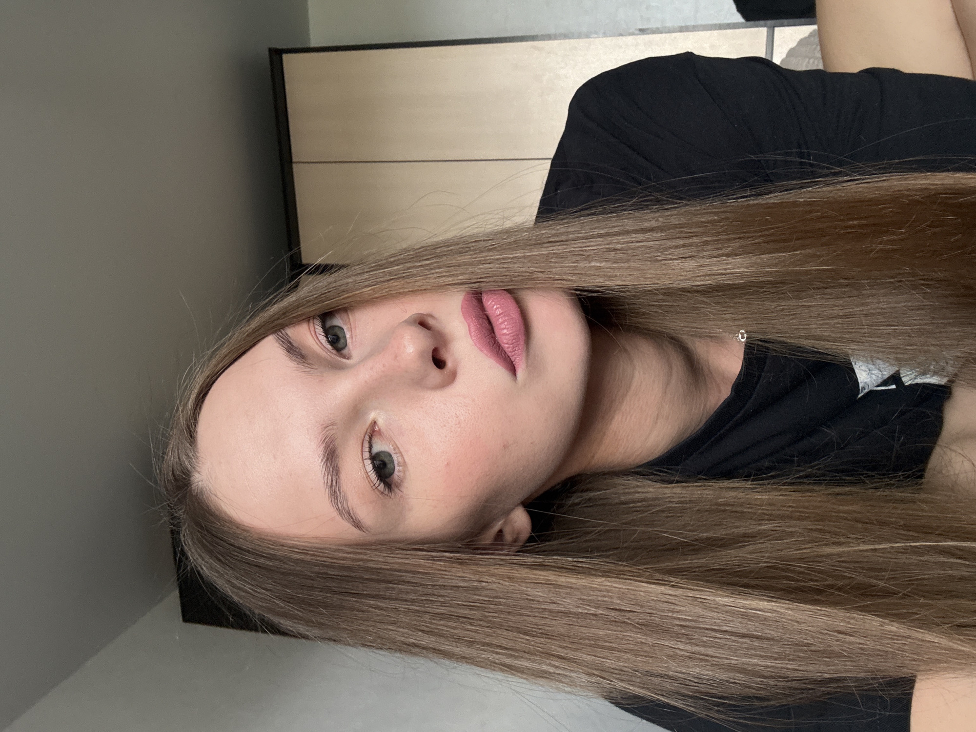 A young woman with long, light brown hair and pale skin is shown from the chest up in a close-up, slightly angled shot. She is wearing a black t-shirt. Her hair is straight and falls around her face and shoulders. She is looking towards the camera with a neutral expression. Her lips are slightly parted and appear to have light pink lipstick on. Her eyes are open, and her gaze is directed slightly upwards. The background consists of a blurred wall and a piece of furniture with a light wood grain. The lighting suggests an indoor setting, possibly during the daytime. There is no visible text in the image. The image is oriented sideways.