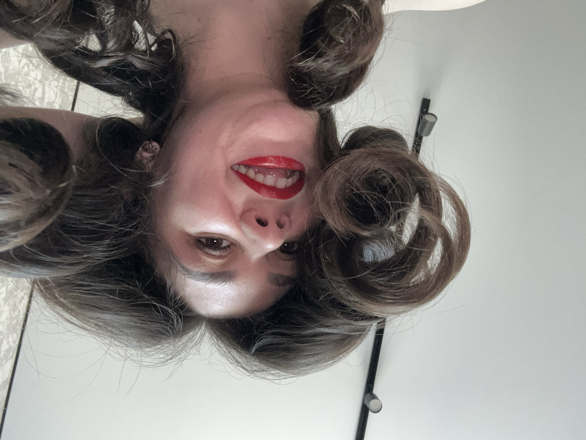 A photograph features a woman with curly brown hair and red lipstick, taken from a low, inverted angle. Her face is centered in the frame, with the background showing a white wall and a portion of what appears to be a lighting fixture. No information suggests the image was taken in Chaikovsky, Russia.