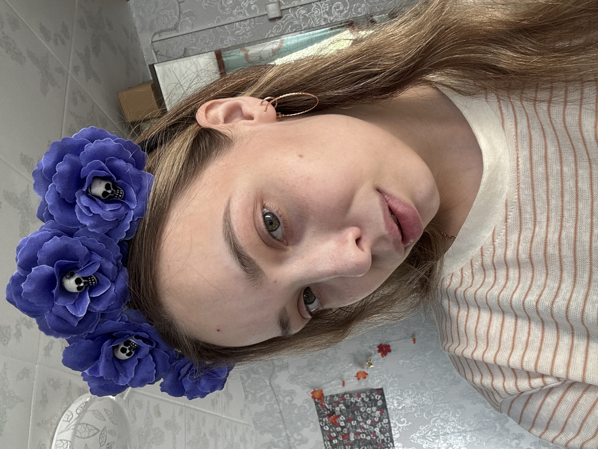 A close-up, angled shot depicts a fair-skinned individual with light brown hair. They wear a crown of blue fabric flowers, each containing a small, white skull in the center. The person has gold hoop earrings. They wear a white top with thin, orange vertical stripes. The background includes a tiled wall, a portion of a mirror, and a patterned tablecloth. The location is Кемуль, Russia.