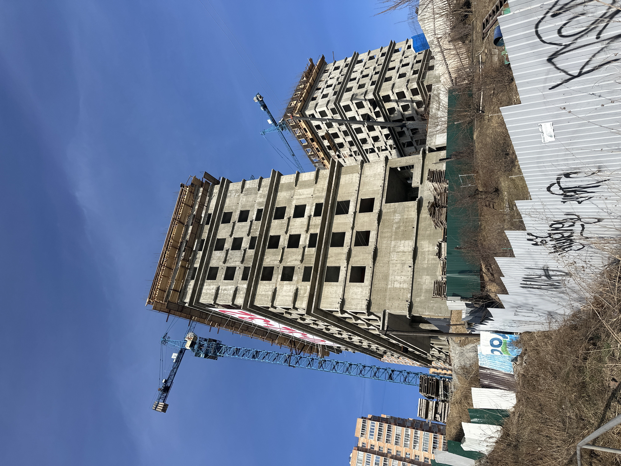 The image, taken in Irkutsk, Russia, depicts an active construction site under a clear, bright blue sky. Two large, multi-story concrete buildings, appearing to be residential or commercial, are the primary subjects. The building closer to the viewer stands on the left, displaying a raw concrete frame with numerous square and rectangular window openings. Its upper levels show visible wooden formwork for ongoing construction. A prominent blue tower crane with its jib extending towards the right is positioned in front of this building.
Further back and to the right, another similar concrete structure is under construction, appearing taller and with a more complete facade of repetitive square openings. A second, smaller tower crane is visible near its upper sections. The clear, sunny weather casts distinct shadows, highlighting the architectural details and the repetitive patterns of the structures.
In the immediate foreground, a corrugated metal fence, partly grey and covered with black graffiti, and partly green, encloses the construction area. Beyond the fence, dry, sparse vegetation, consisting of bare bushes and weeds, suggests a late winter or early spring setting. In the distant background, another completed multi-story brick building with numerous windows is visible, indicating an urban environment.
Visible text includes illegible black graffiti on the grey fence panels, a large red and white banner on the side of the closer concrete building displaying the Russian word "БЕЗ" (BEZ - "WITHOUT"), and a small blue and white poster on the green fence section. The scene captures a moment of active urban development.