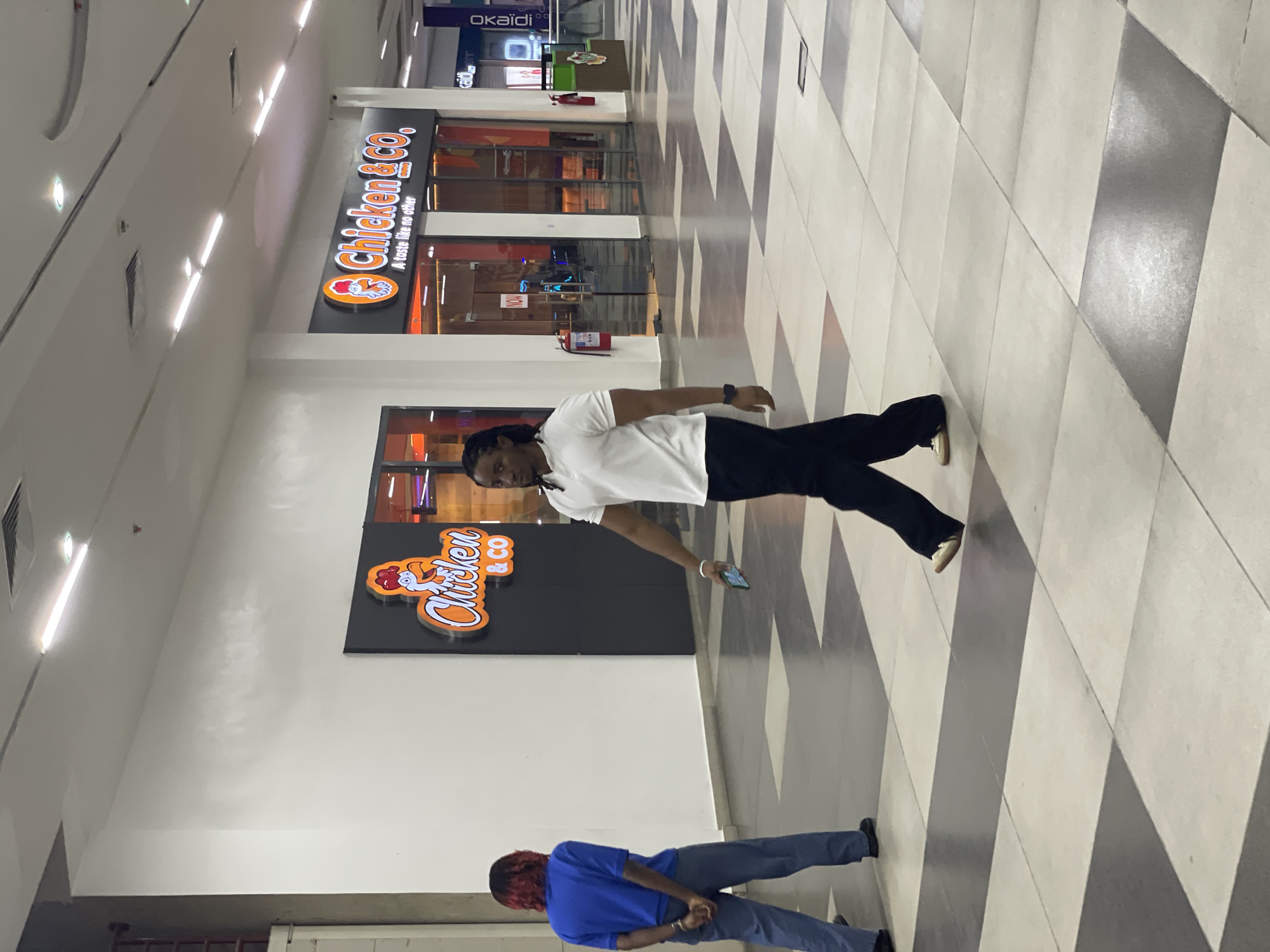 In a brightly lit indoor shopping mall hallway, two individuals are captured walking. The individual on the left, with reddish-brown hair, is wearing a blue short-sleeved shirt and grey trousers, facing away from the camera and holding their hands behind their back. The individual on the right, with dark braided hair, is wearing a white polo shirt and black trousers, walking towards the right side of the frame, looking at their phone which they hold in their right hand.
In the background, the storefronts of two restaurants are visible. The "Chicken & Co." restaurant has illuminated signage featuring a cartoon chicken and the text "A taste like no other." The restaurant appears to be open, with interior lighting and glass doors. Fire extinguishers are mounted on the walls near both establishments. The floor is tiled in a repeating pattern of light and dark grey squares. The overall environment suggests a commercial shopping area.